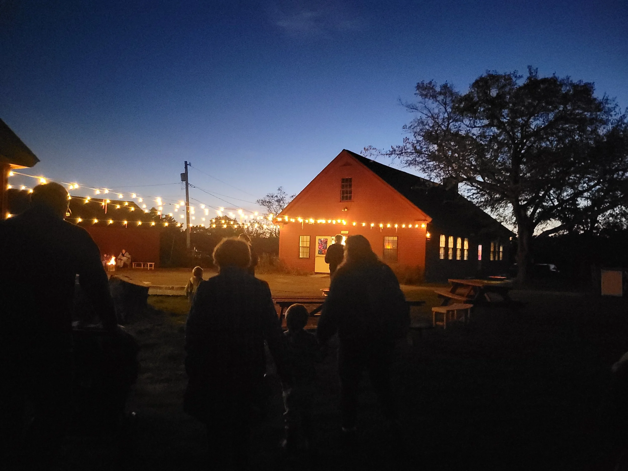 Outdoor gathering at dusk with people standing and sitting under string lights near a red building with large windows, trees, and a clear evening sky.