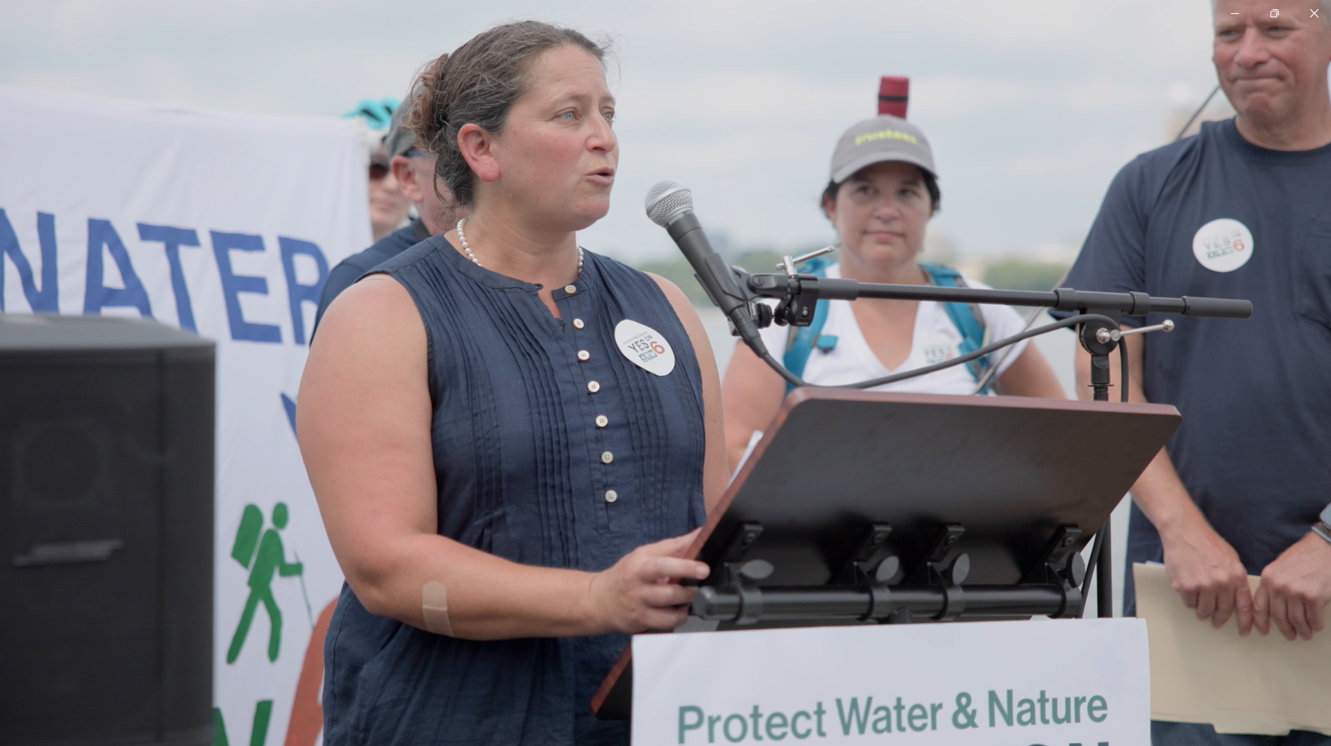 A woman speaking at a podium during a protest, with a banner behind her and several people standing nearby. The banner reads 'Protect Water & Nature'.