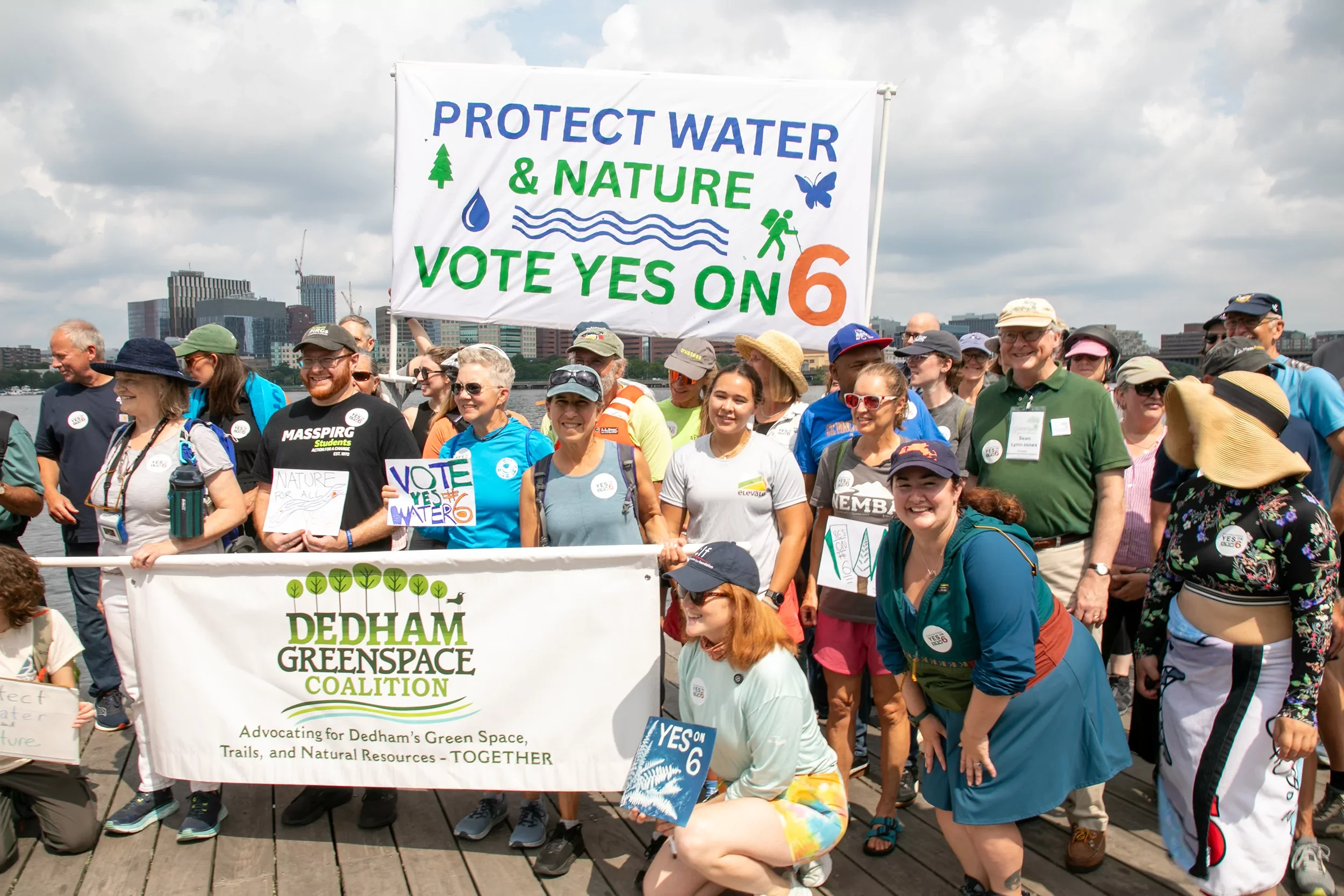 Group of people participating in a rally, holding a large banner that reads 'Protect Water & Nature, Vote Yes on 6' during daytime, with city buildings and cloudy sky in the background.