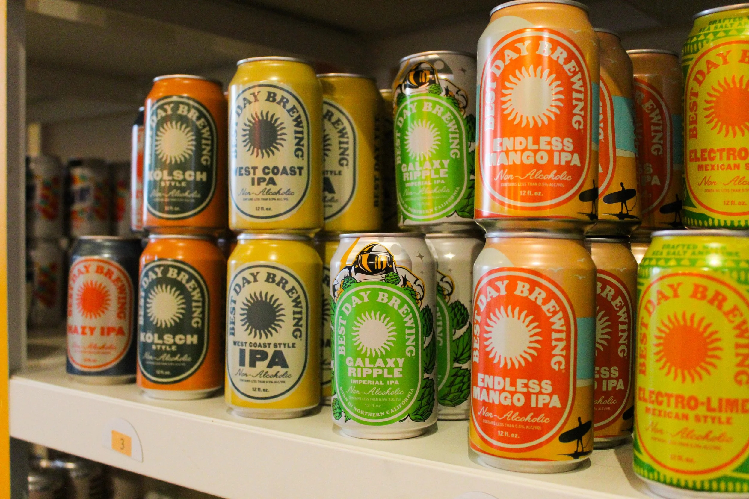 Shelf displaying various non-alcoholic craft beer cans with colorful labels, including 'Endless Mango IPA' and 'Galaxy Rippie' on a store shelf. marigold bottle shop photo by Atika Goble branding and product photographer in minneapolis atikagoble.com