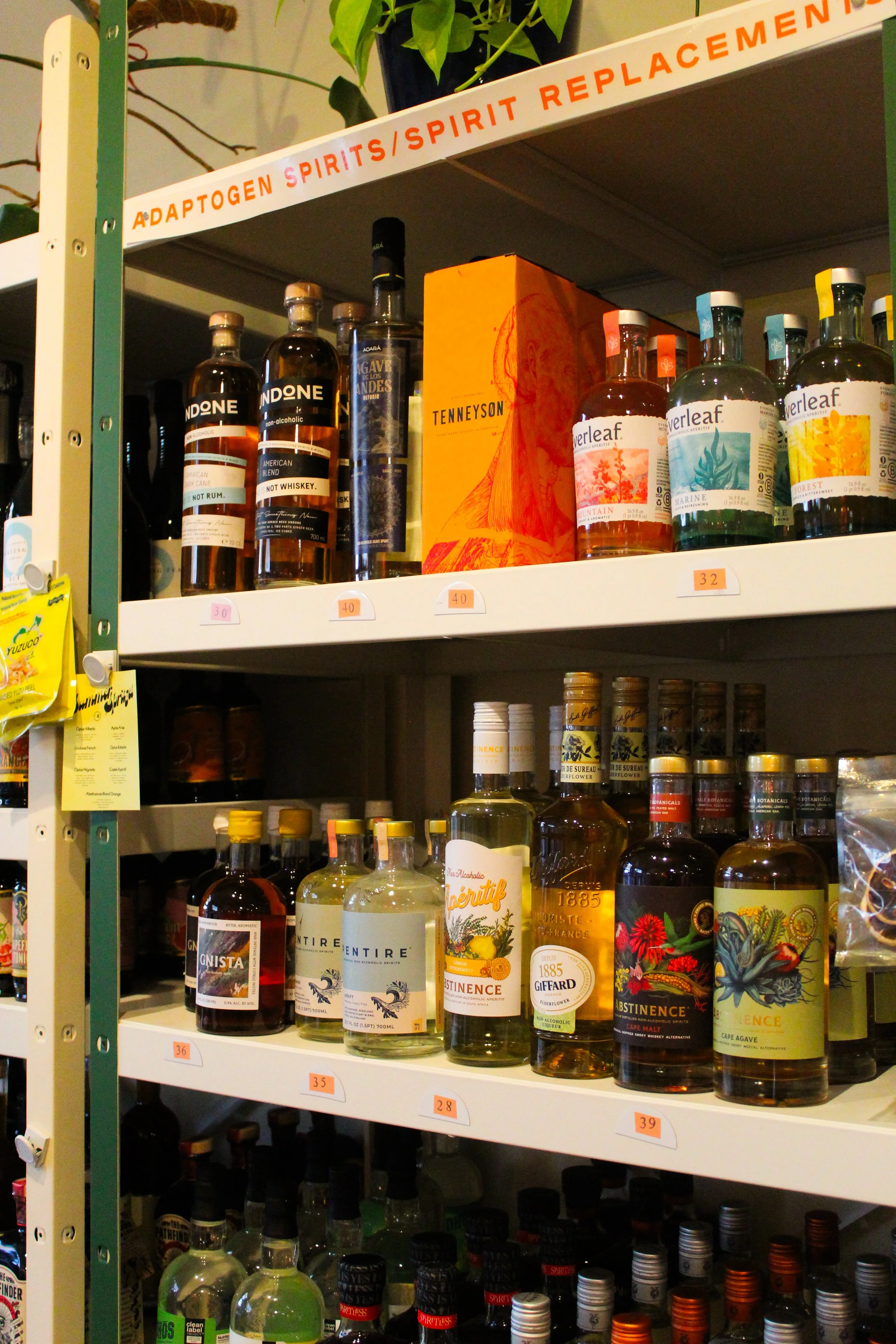 Shelf with various bottles of spirits including whiskey, flavored liqueurs, and botanical spirits, some labeled with brand names such as Verleaf and Notre Alcoholic. A plant is visible on the top shelf. The shelf has labels with prices and a sign tha