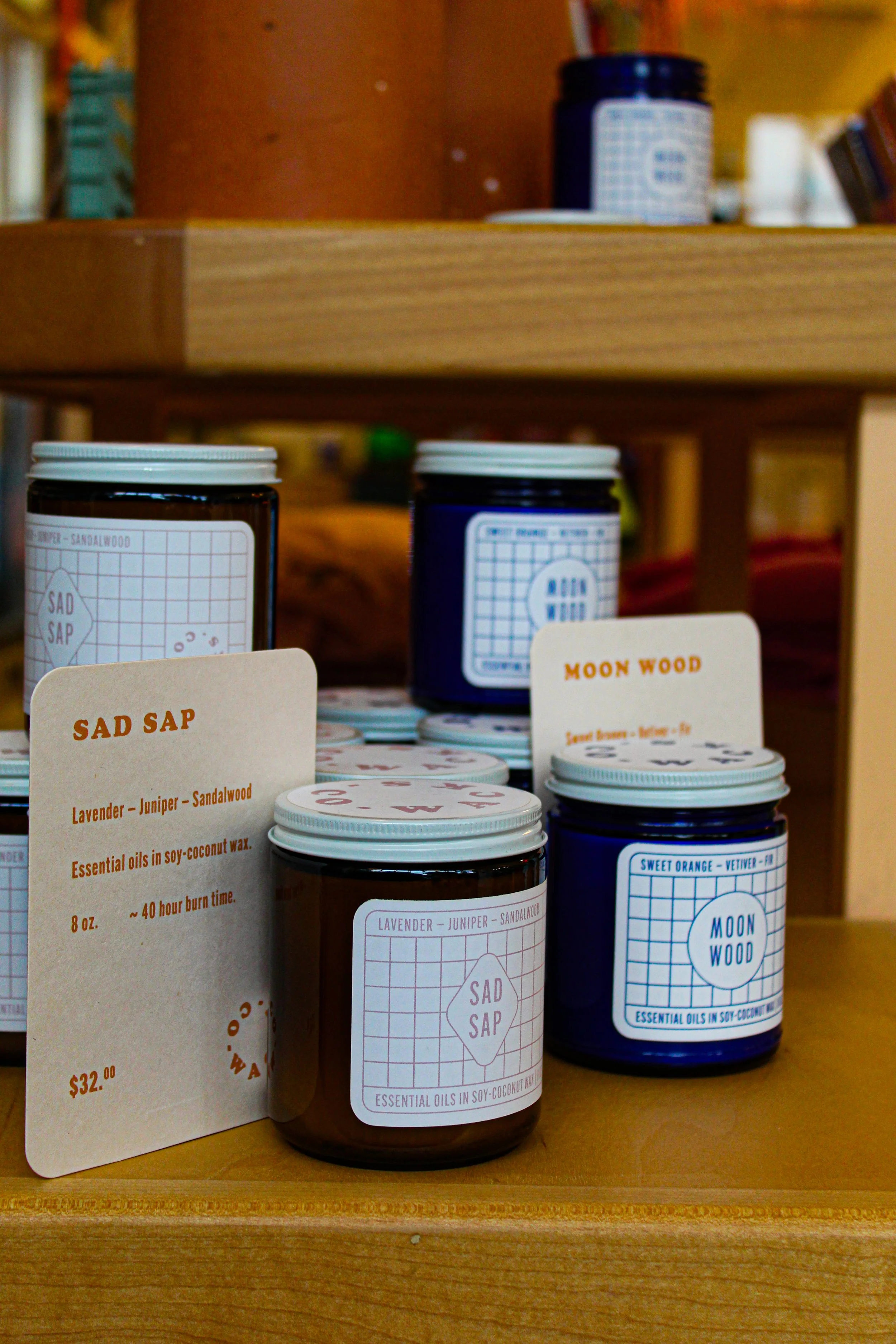 Two jars of scented candles on a wooden display shelf with price tags and labels, one labeled "Sad Sap" with lavender, juniper, and sandalwood, and the other labeled "Moon Wood" with sweet orange scent. photo by Atika Goble branding and product photo