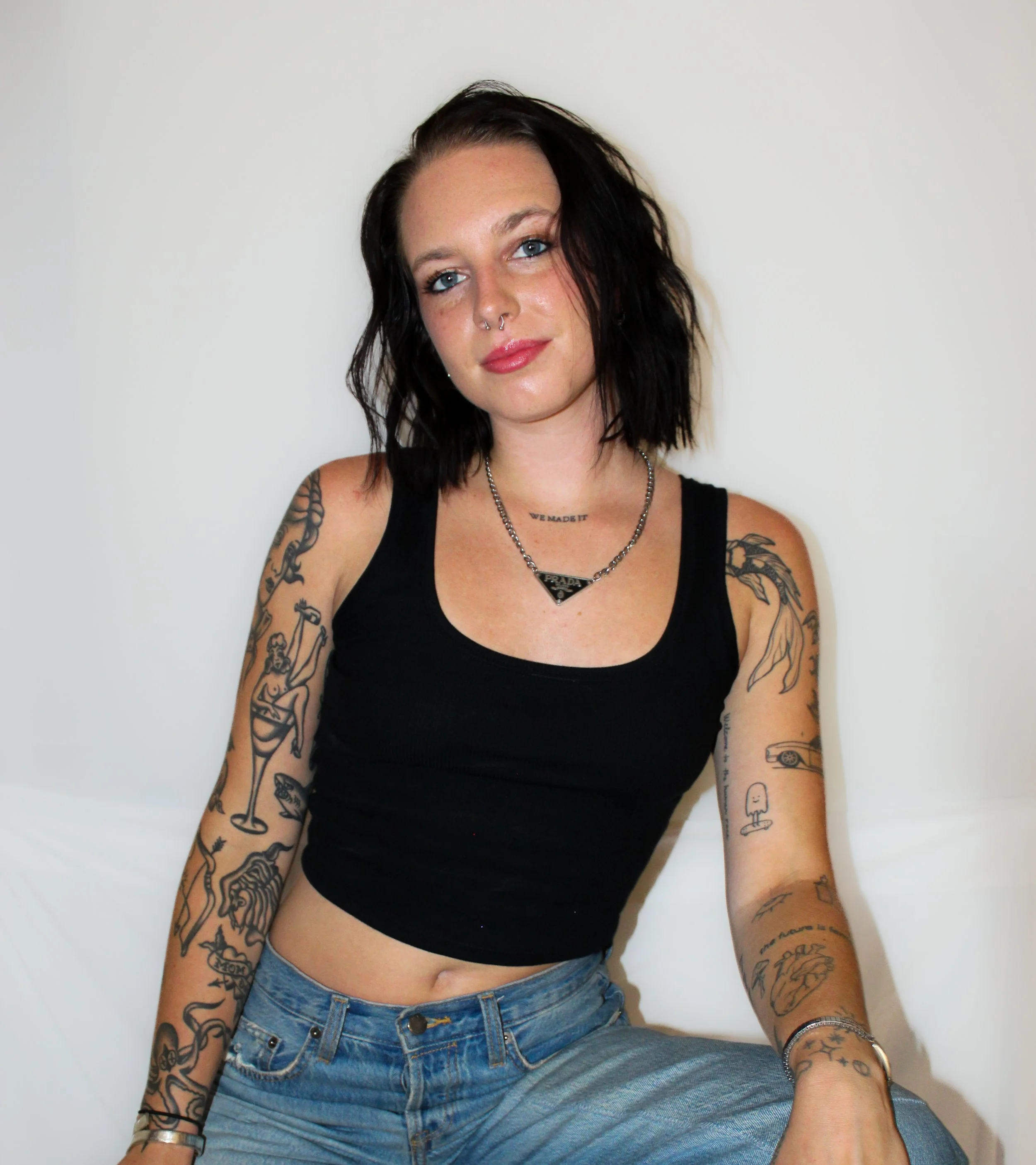A young woman with black wavy hair, tattoos on both arms, blue eyes, and wearing a black crop top and jeans, sitting against a plain white wall.