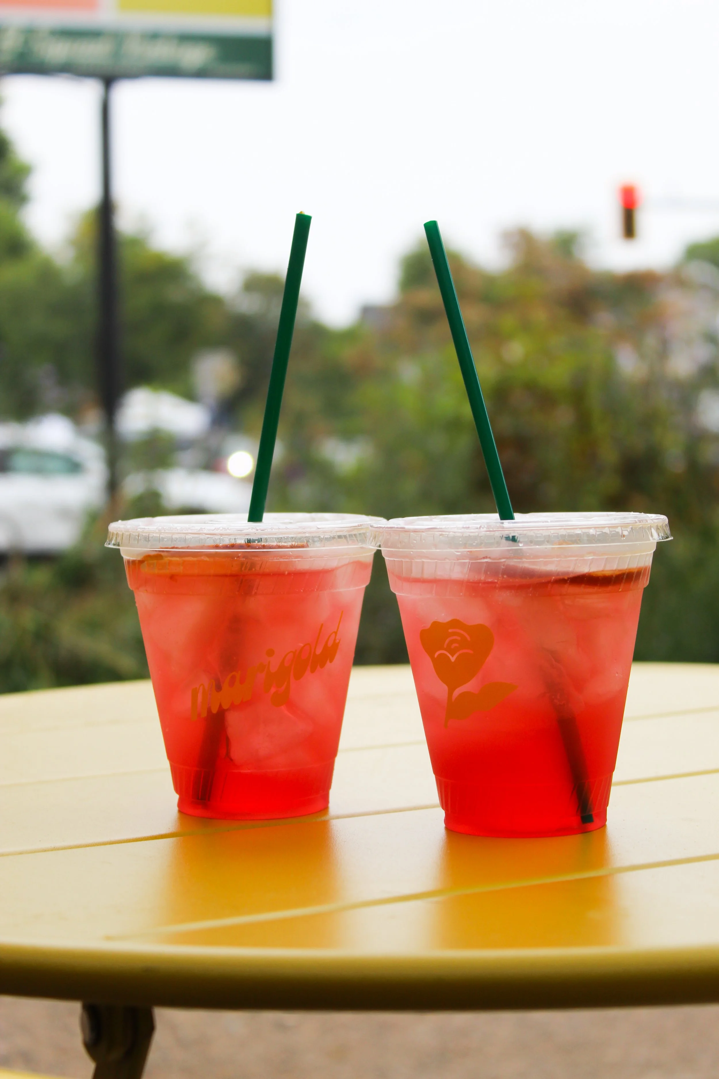 Two clear cups filled with a reddish-pink beverage and ice, with green straws, sitting on a yellow table.