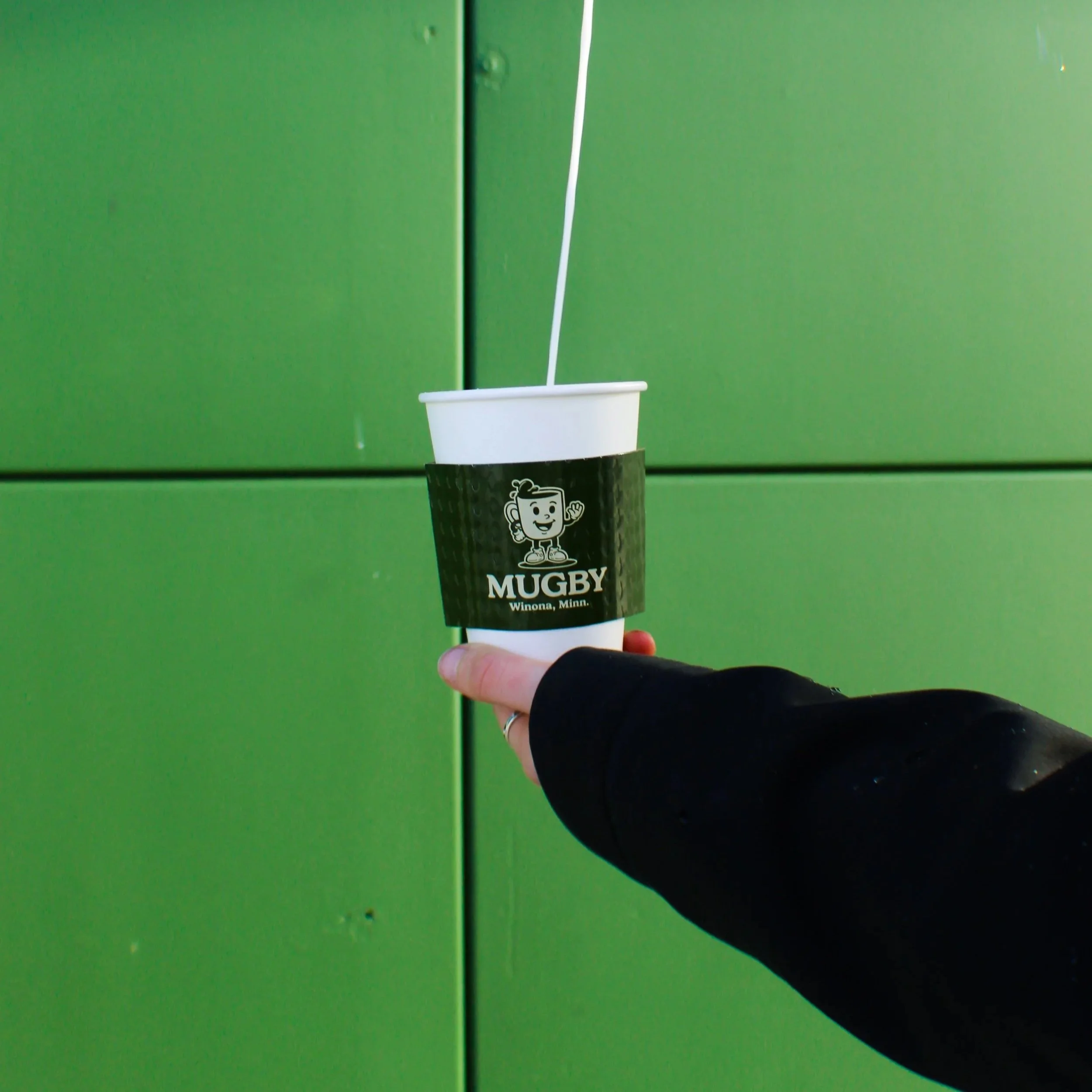 A hand holding a cup with the logo of Mugby, a coffee shop from Winona, Minnesota, against a green background.