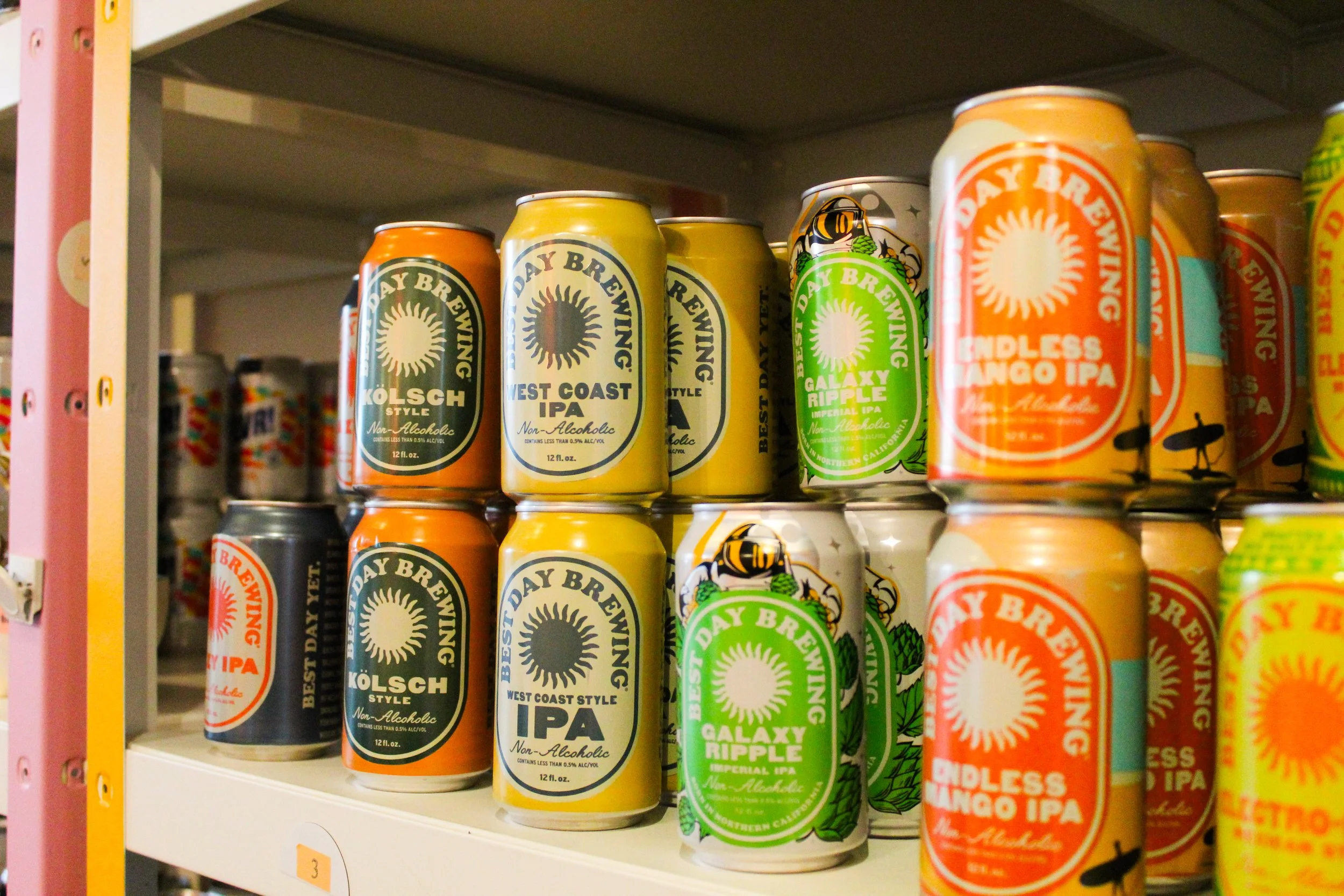 Shelf displaying various canned craft beers, including Kolsch Style, West Coast IPA, Galaxy Ripple Imperial IPA, and Endless Mango IPA, with colorful labels and designs.