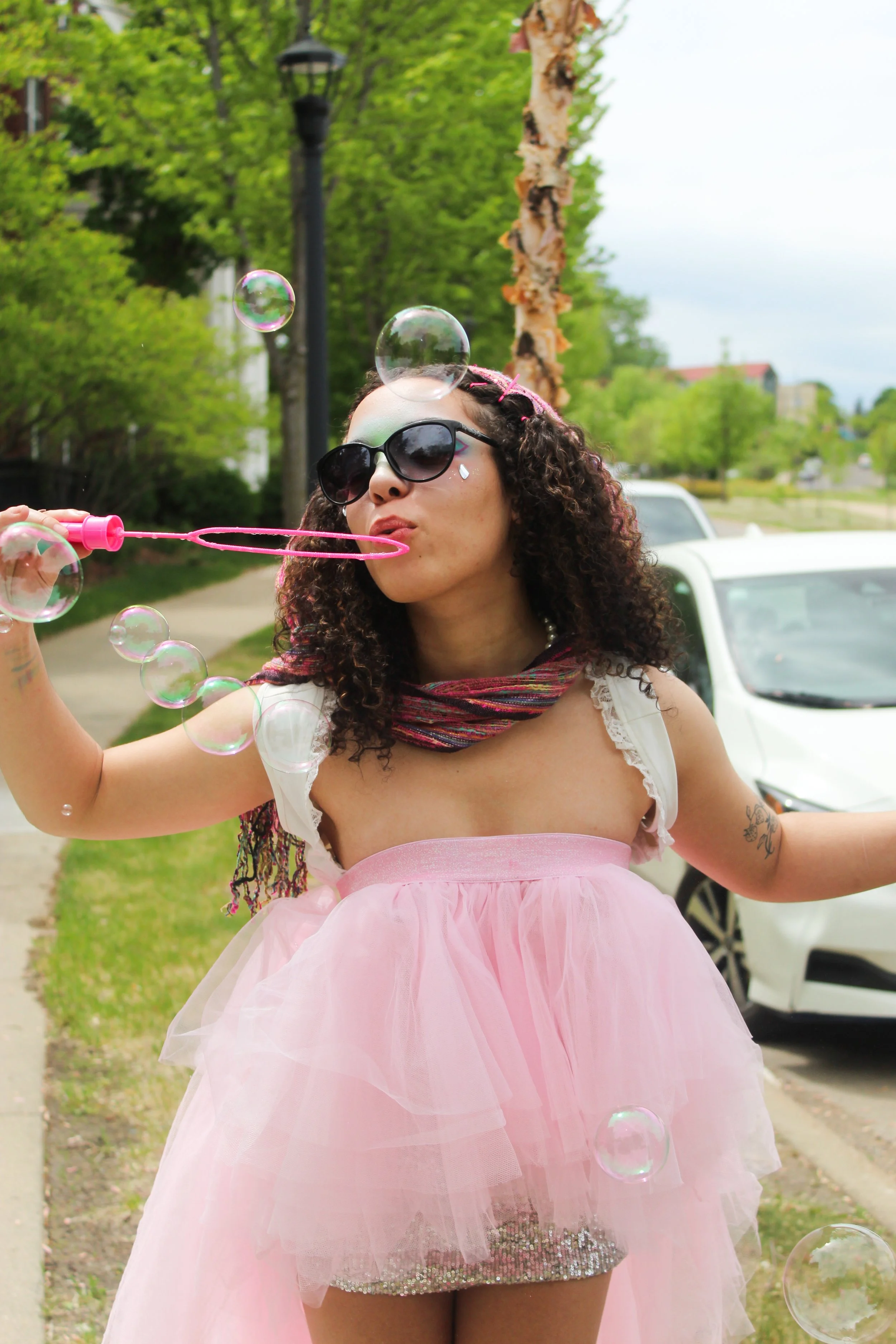 A woman with curly hair wearing dark sunglasses, a pink dress, and a multicolored scarf blowing bubbles outdoors on a sunny day with trees and cars in the background.