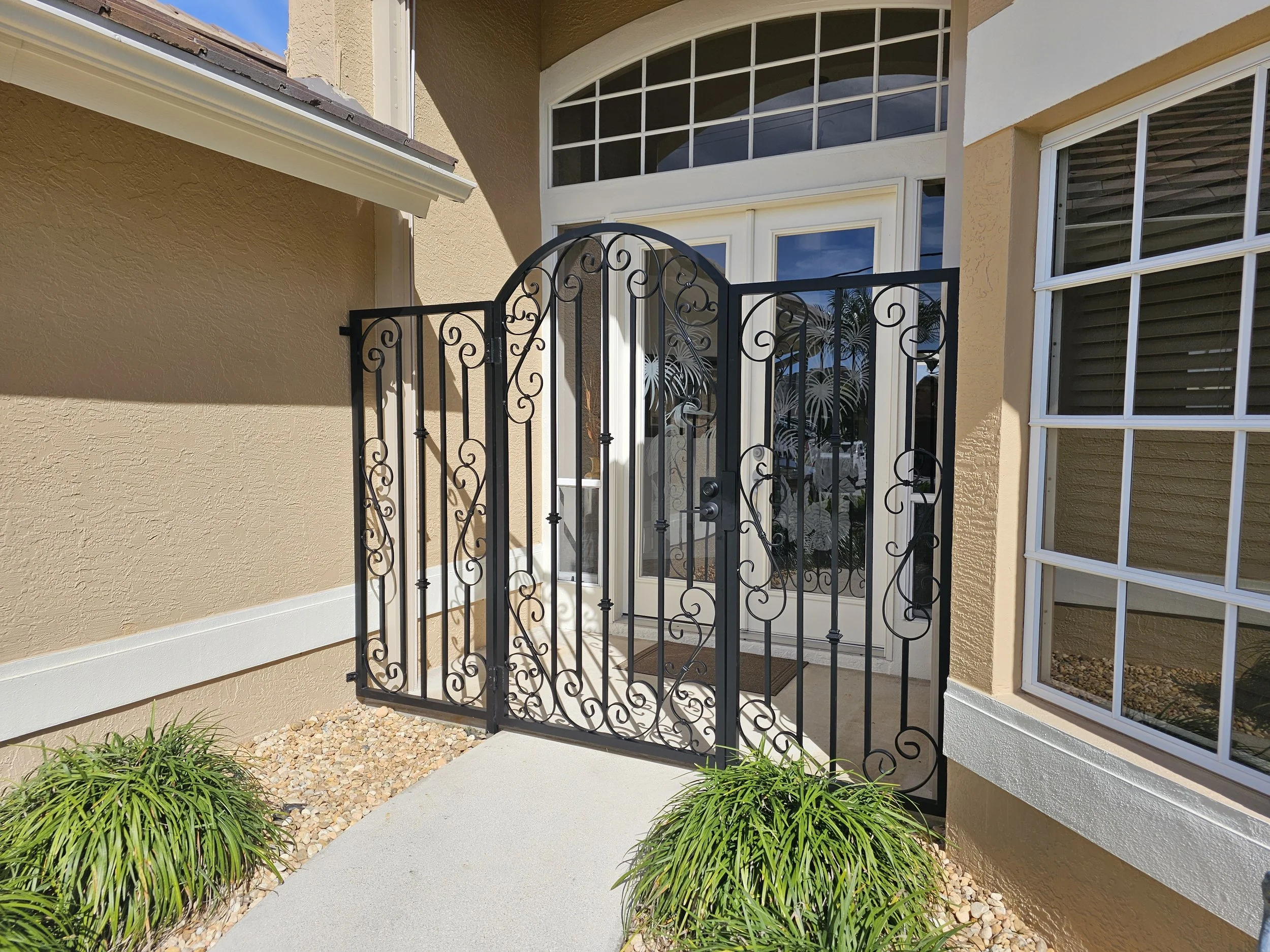 Black decorative aluminum entry gate in front of a front door with large glass panels, part of a house with beige textured walls and white trim, landscaped with green plants and small rocks.