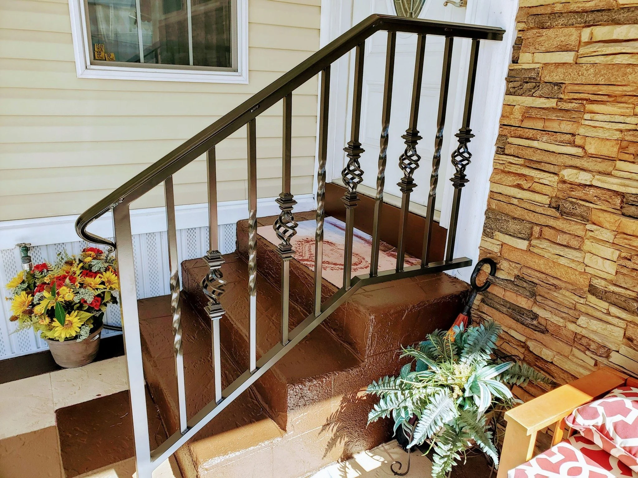 Front porch steps with decorative black metal railings, a potted sunflower and red flowers, green fern-like plant, nearby gardening tools, and a couch with red and white patterned cushions.