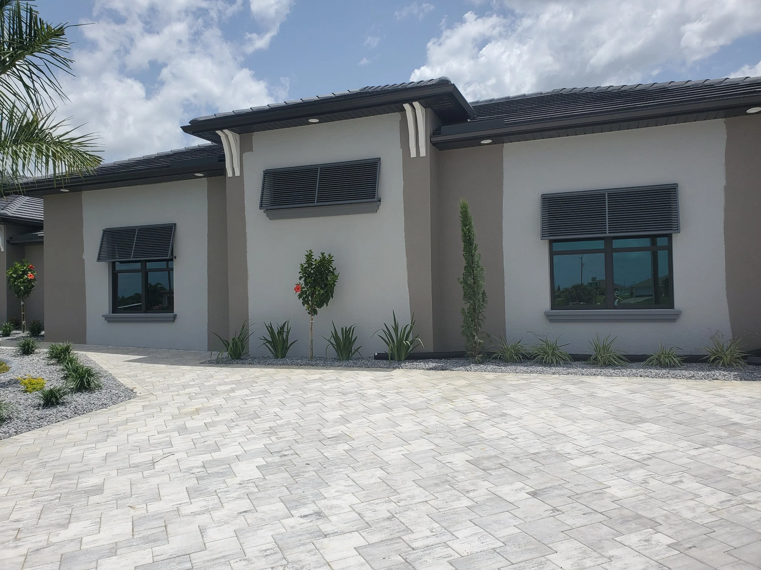 Modern residential building with gray and white exterior,  window louvers, landscaped front yard with small trees and plants, light-colored paved driveway, under a partly cloudy sky.