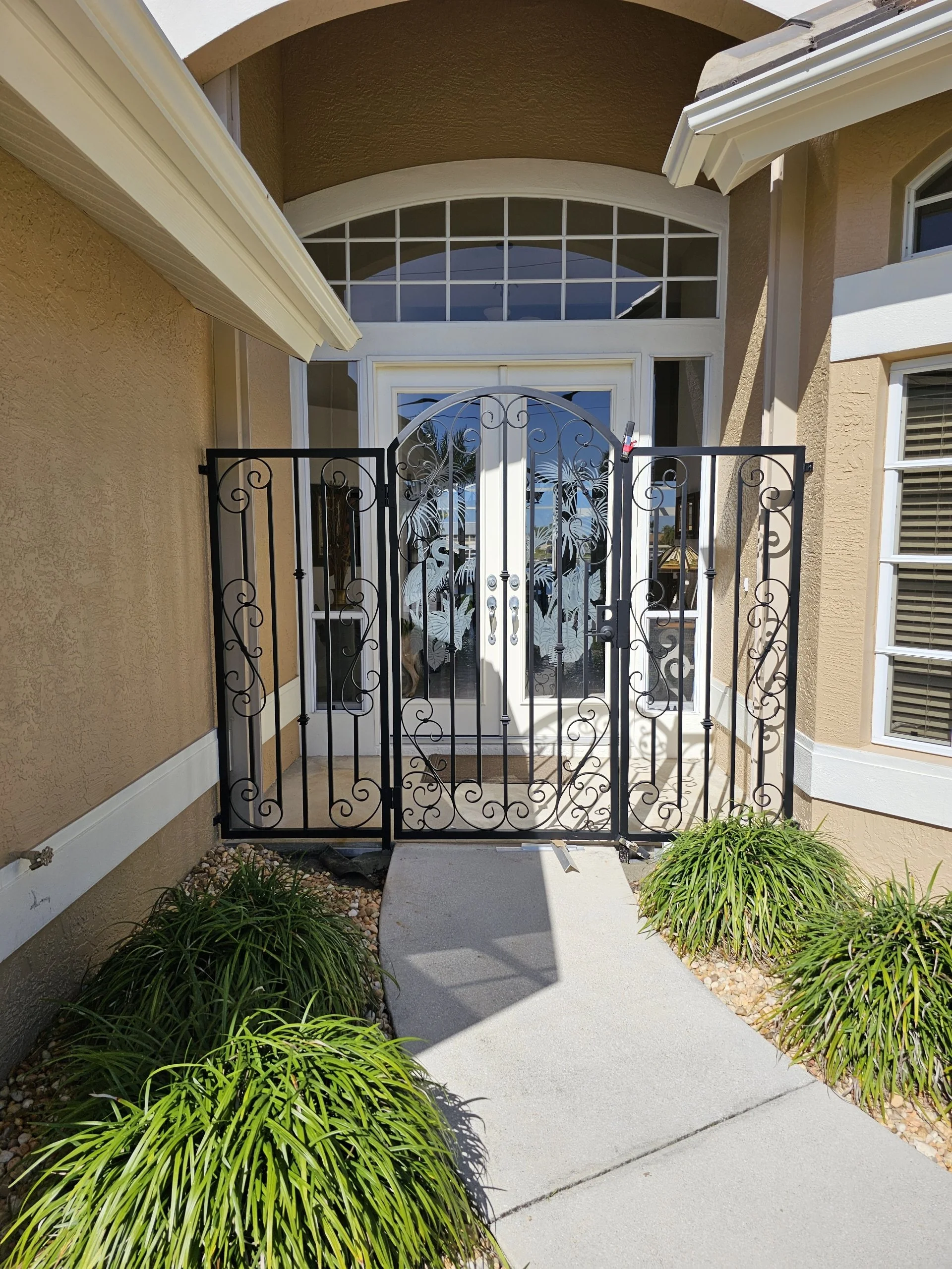 Front entrance of a house with decorative aluminum entry gate, glass double doors, and landscaped bushes.