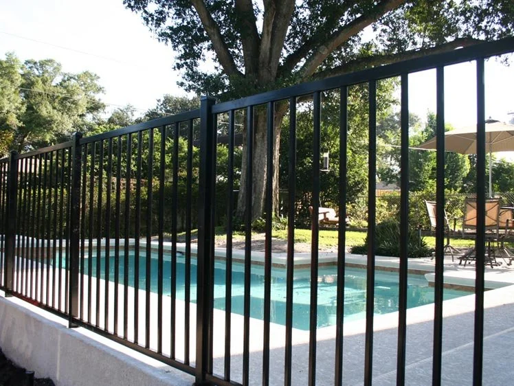 Swimming pool surrounded by a black metal fence with trees and outdoor seating in the background.