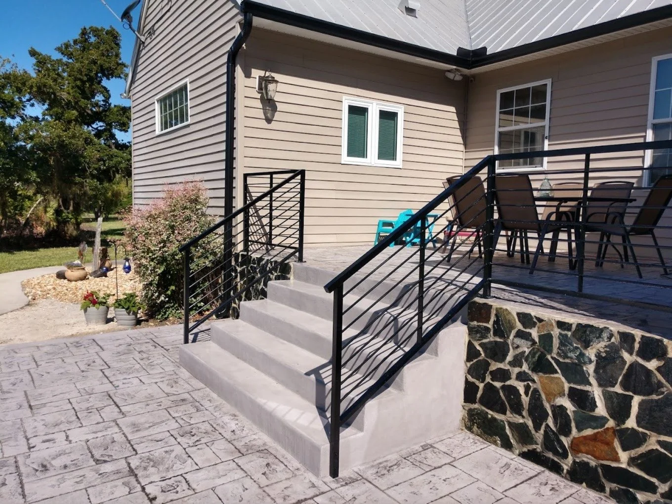 Back porch of a house with stairs, black metal railing, outdoor furniture including four chairs and a table, potted plants, and a landscaped yard with trees.