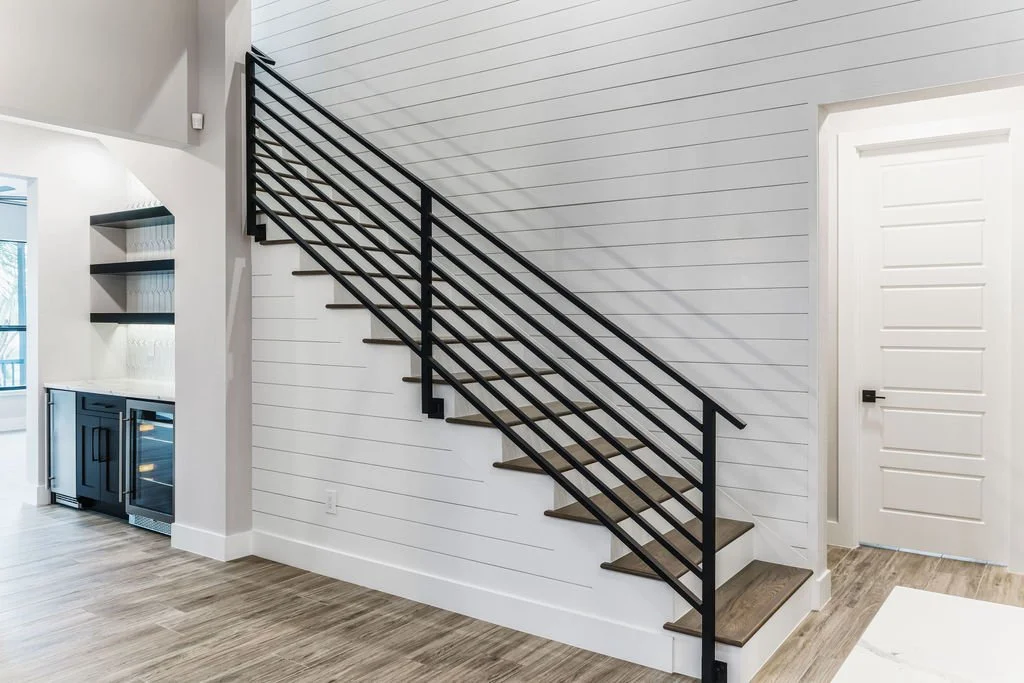 Interior view of a staircase with black metal railing, wooden steps, and white shiplap wall in a modern home.