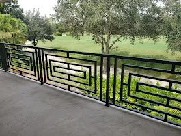 Black metal railing with geometric design on a balcony overlooking a grassy area with trees.