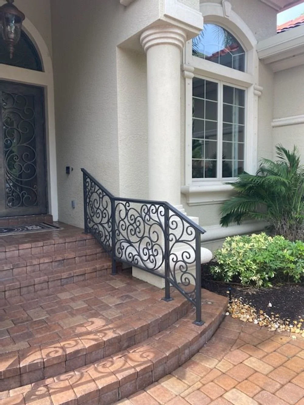 Entrance to a house with brick steps, an ornate black metal handrail, a white textured wall, a large window, and landscaping including green plants.