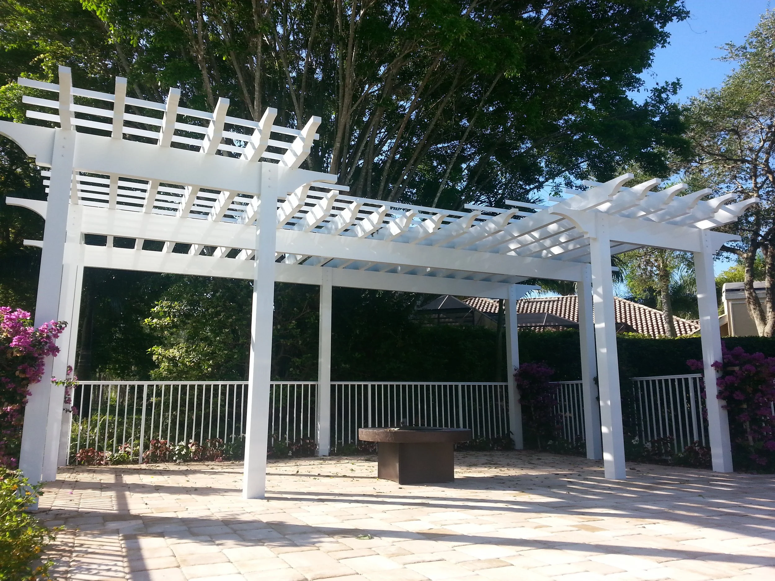 White wooden pergola with lattice roof in a garden, surrounded by flowering bushes and trees, with a paved stone patio and fencing.