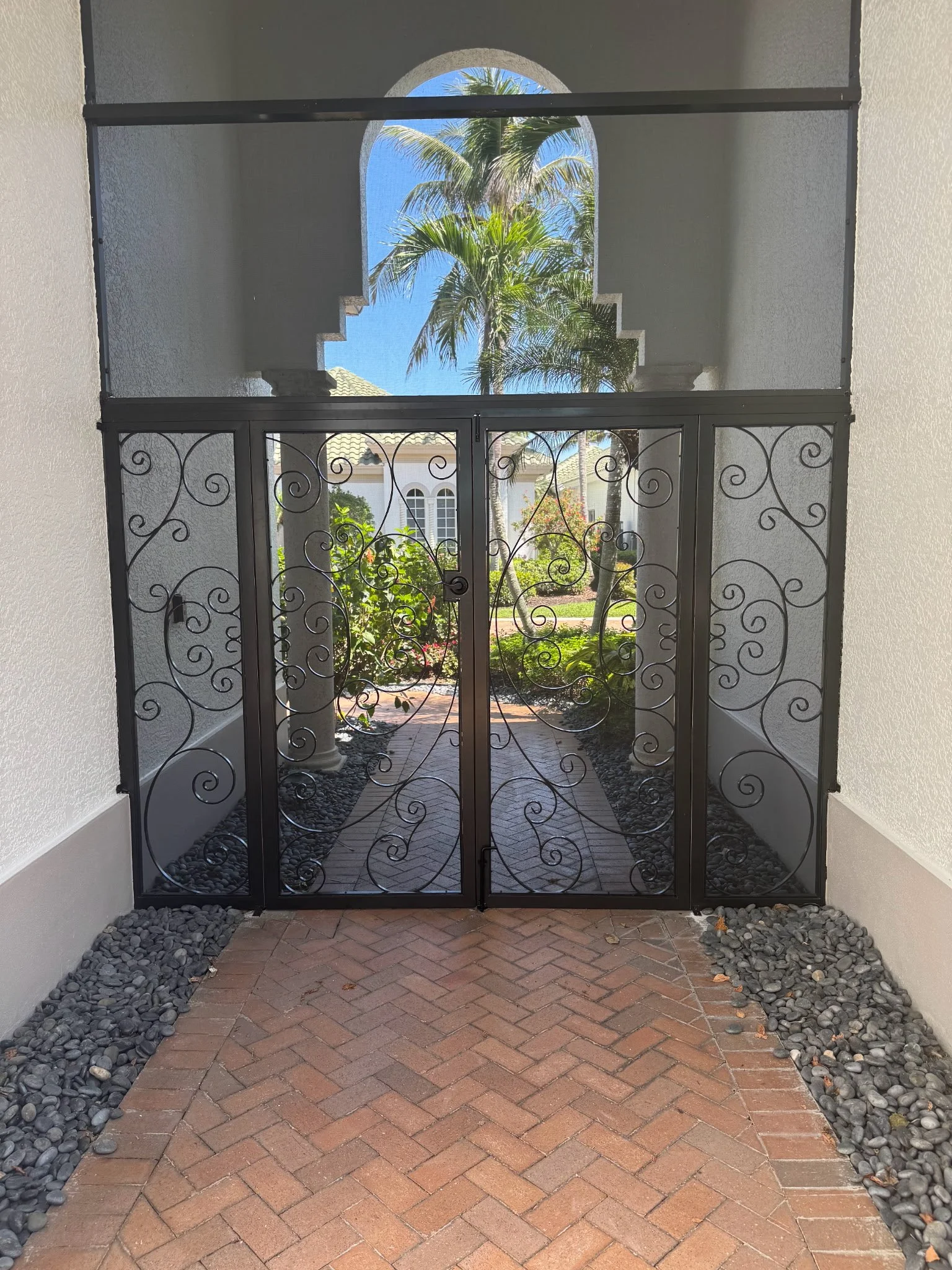 Decorative black aluminum gate leads to a garden with palm trees and a pathway, seen through a screened entrance with an arched window above.