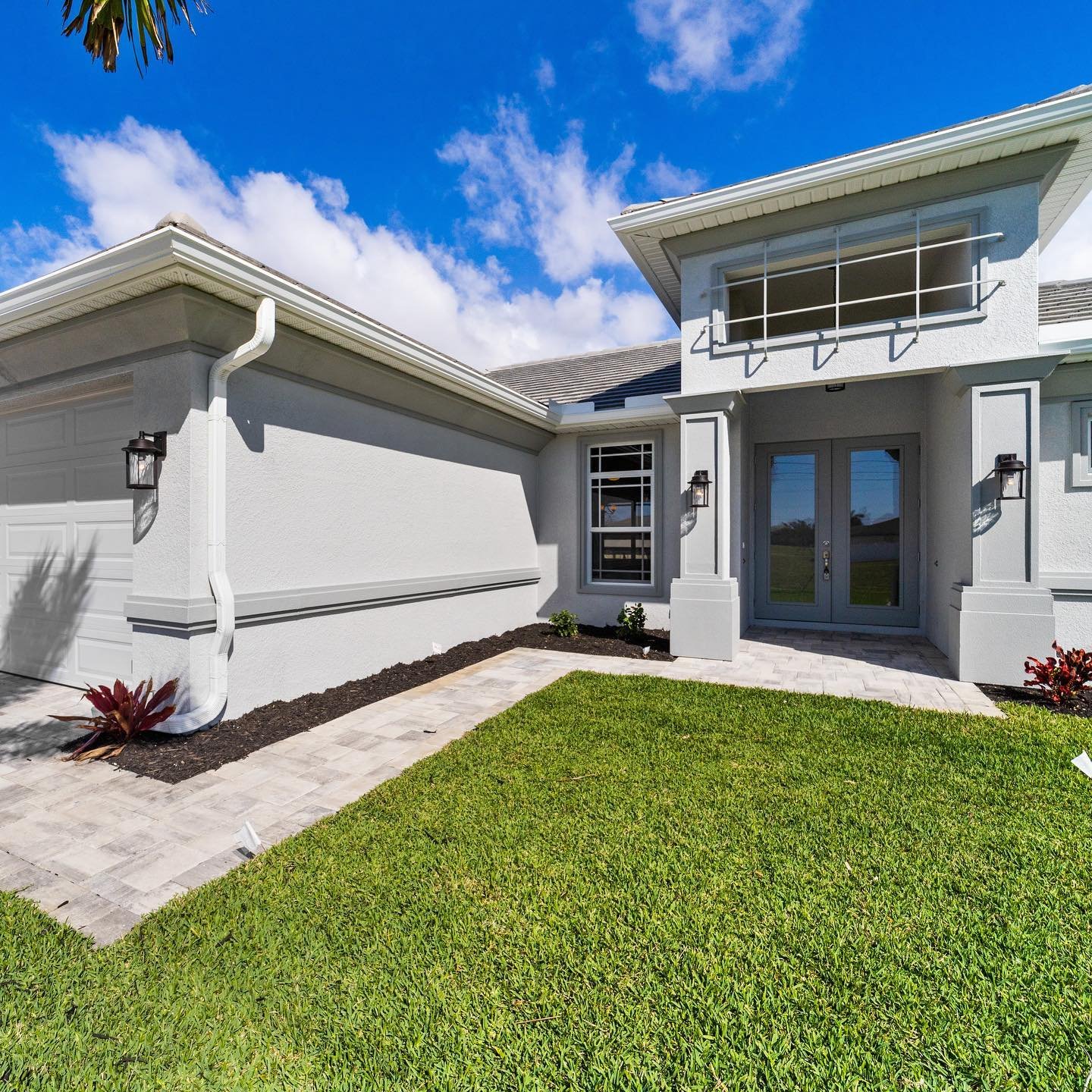 Modern house with gray exterior, deco grills, green lawn, paved walkway, and front door on a sunny day with blue sky and white clouds.