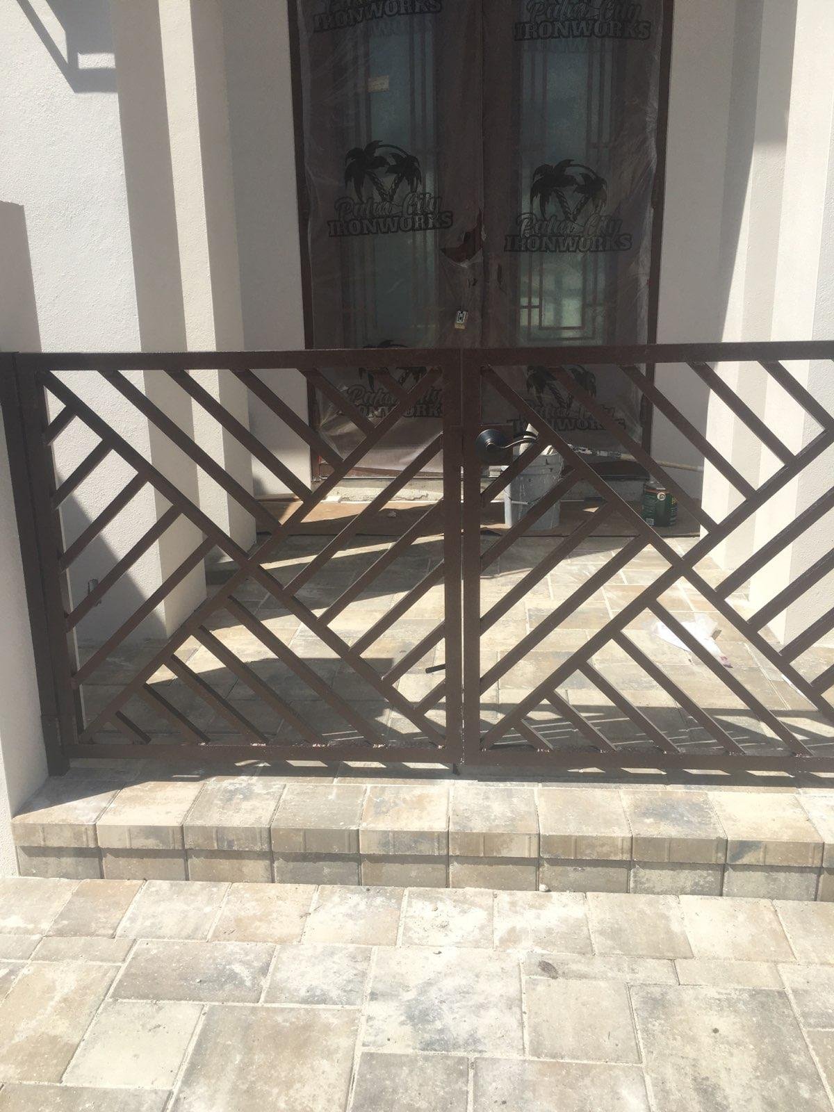 A metal gate with a diagonal crisscross pattern in front of a sliding glass door with a plastic protective covering, on a porch with stone tile flooring.