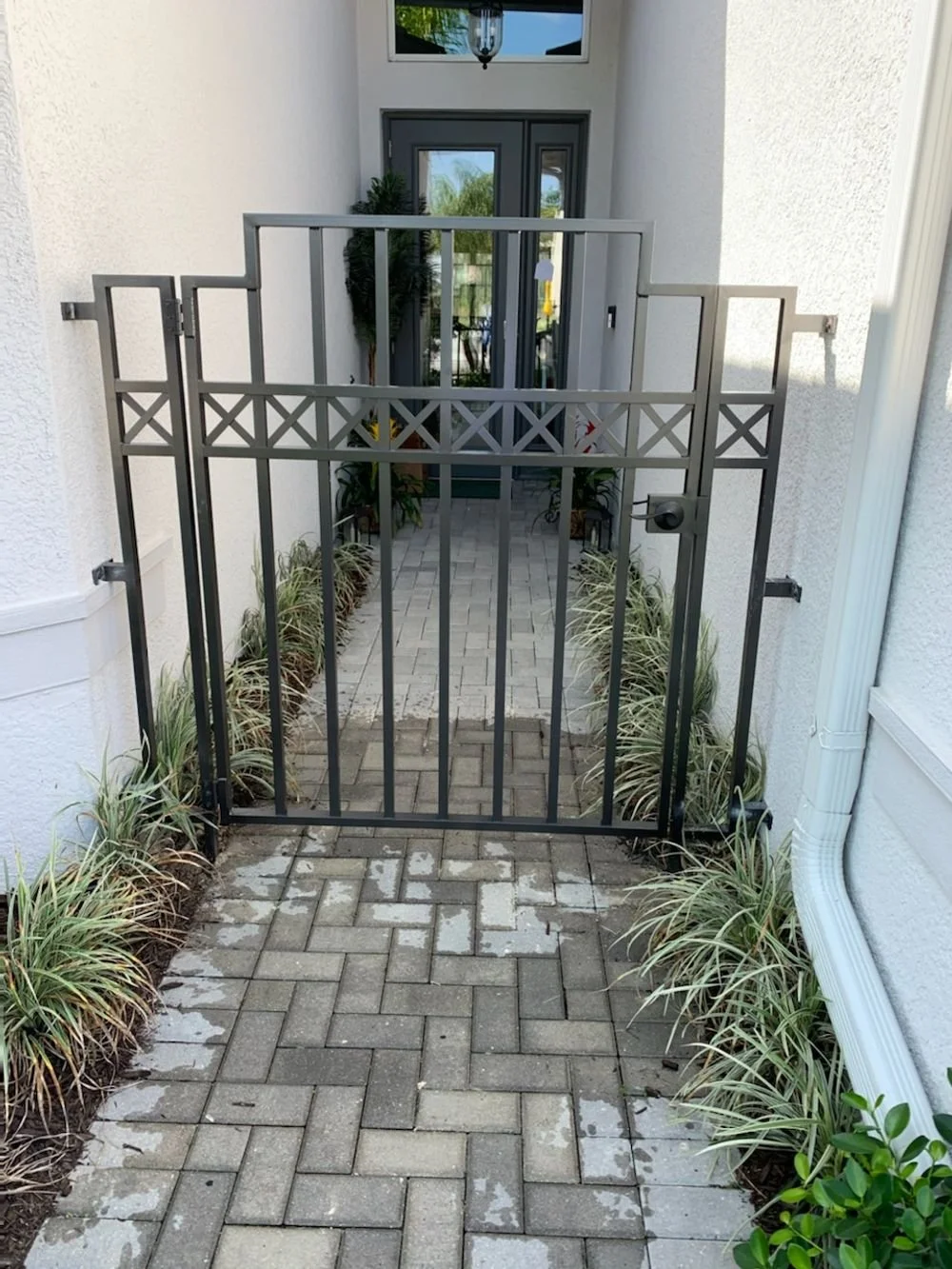 A small gated entryway leading to a front door with potted plants, decorative plants along the sides, and a brick pathway.