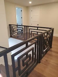 Interior view of a staircase with black metal railing featuring geometric square designs, hardwood flooring, and plain white walls.