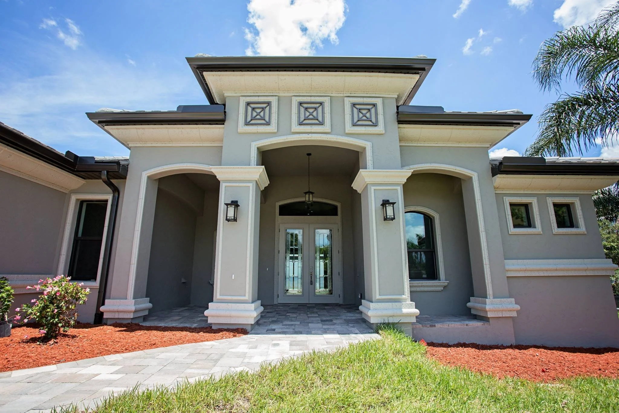 Front view of a modern house with a deco grills, covered entrance, tall windows, and palm trees on the side under a partly cloudy sky.