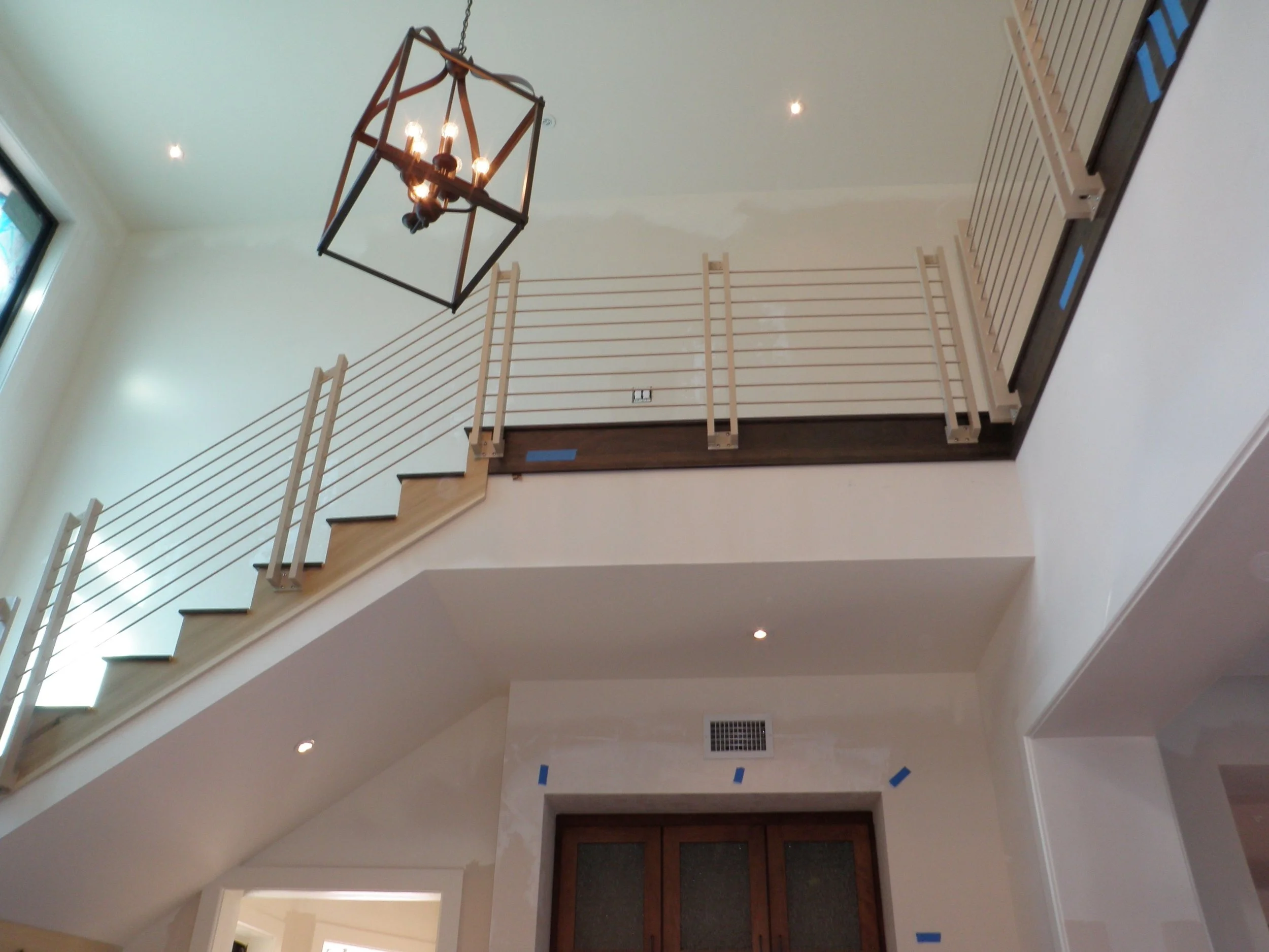 Interior view of a two-story home with a staircase and railing, with a chandelier hanging from the ceiling and various blue painter's tape on the walls and trim, indicating ongoing renovation or painting work.