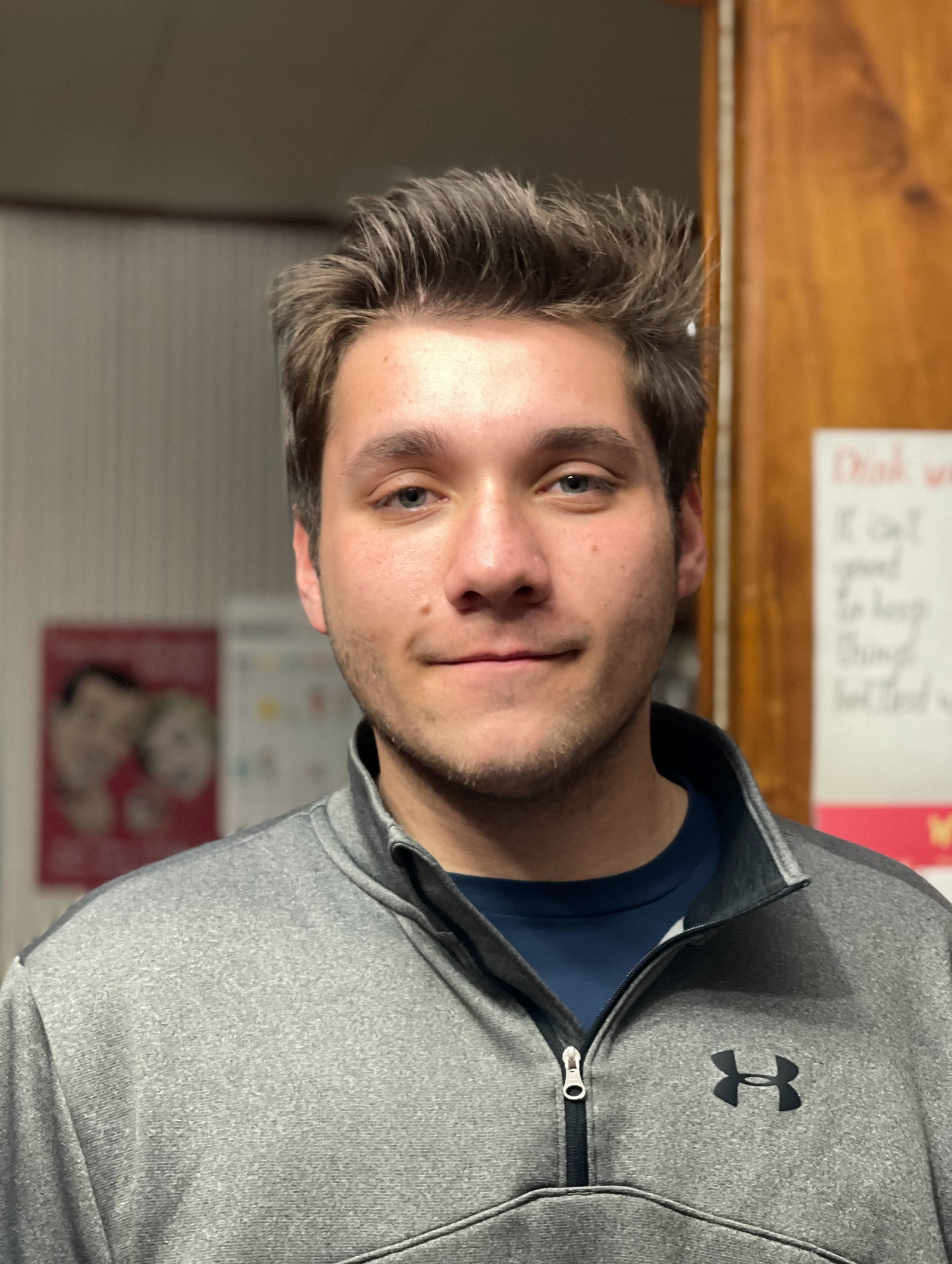 A person wearing a gray Under Armour pullover standing indoors with posters on the wall behind.