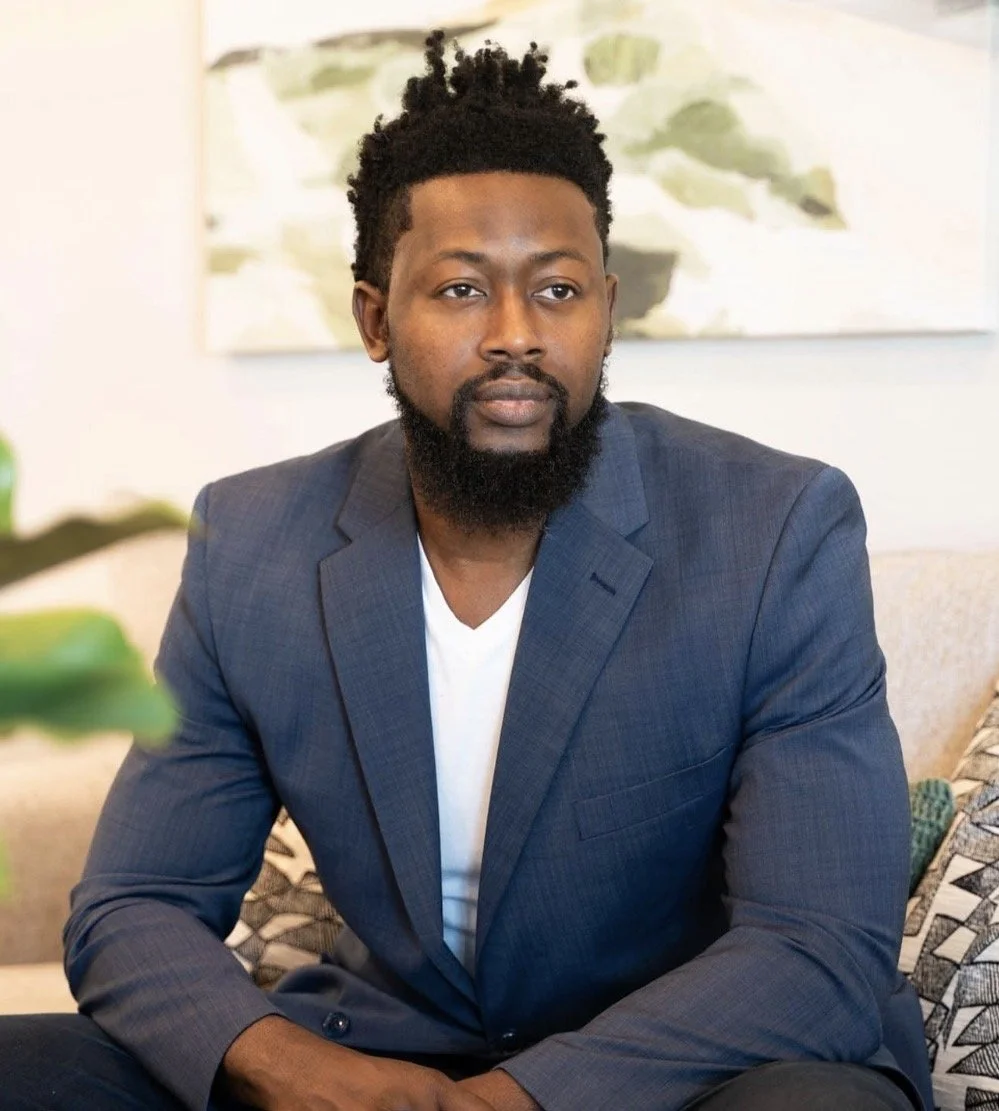 A man with a beard and curly hair, wearing a gray suit jacket and white shirt, sitting on a beige sofa in a room with abstract art on the wall.
