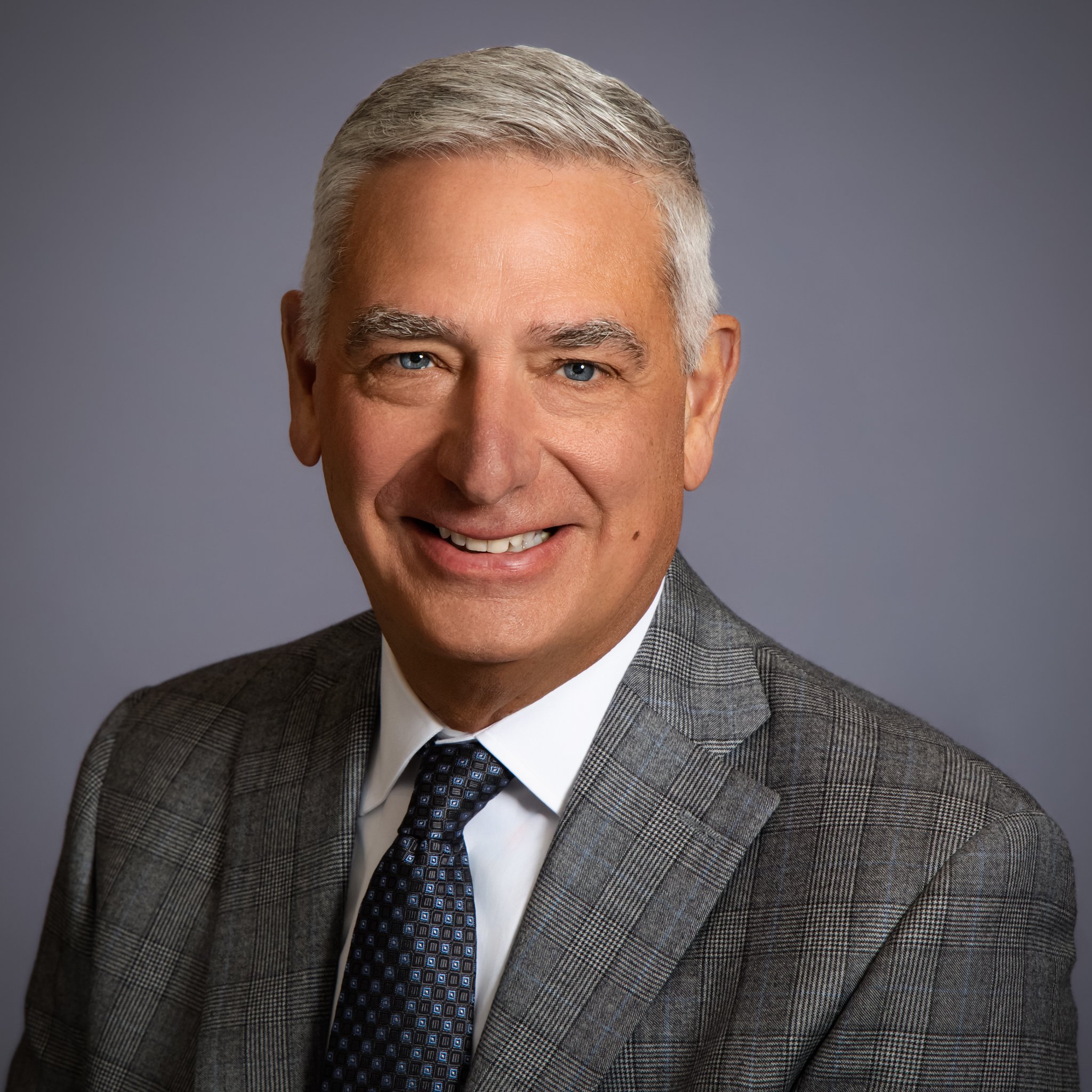 Professional headshot of a middle-aged man with gray hair, light skin, blue eyes, dressed in a gray checkered suit, white shirt, and patterned dark tie, smiling against a plain gray background.