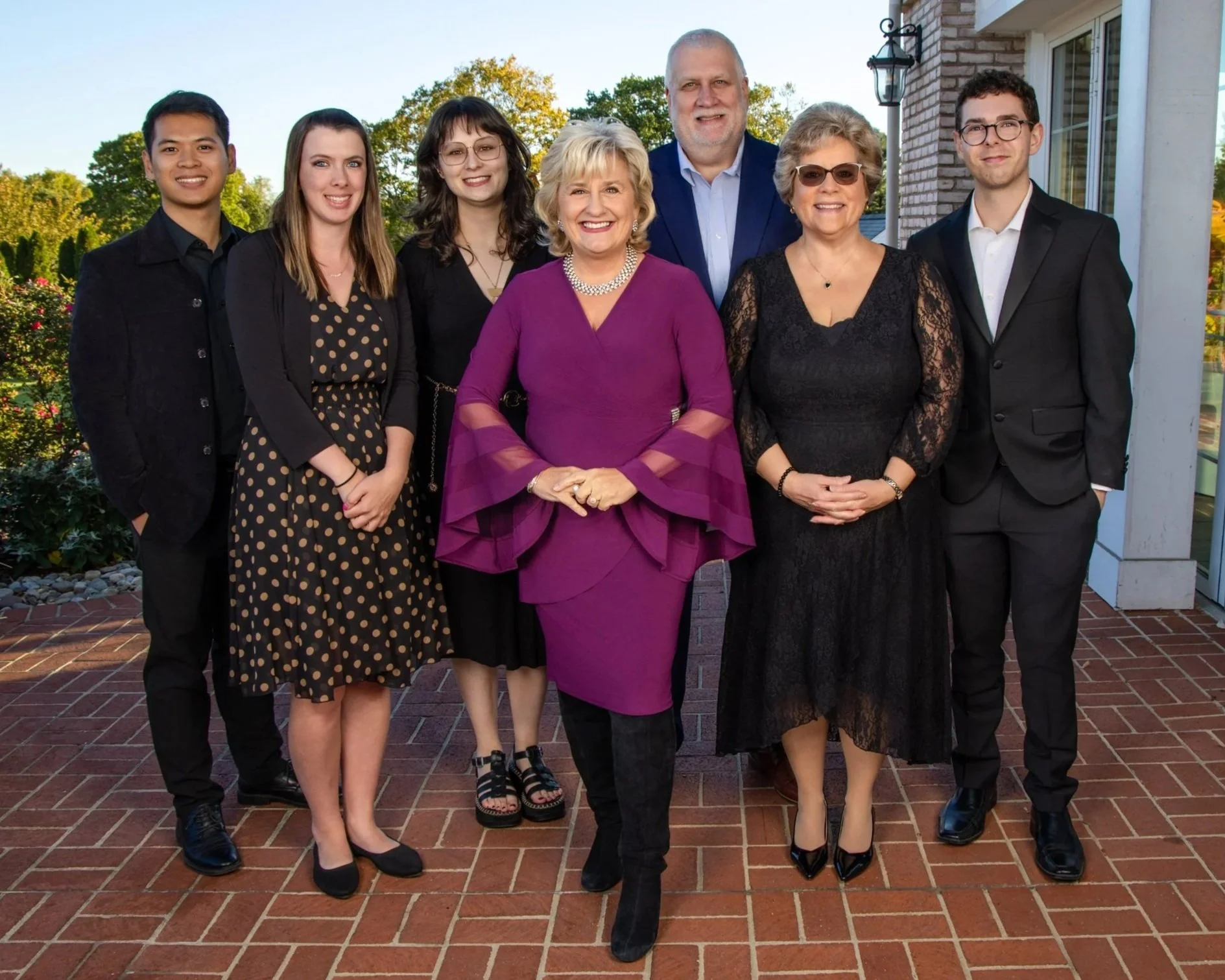 A group of nine people at Gala, including seven women and two men, standing outdoors on a brick patio in front of a house with trees in the background. They are dressed in formal attire and smiling at the camera.