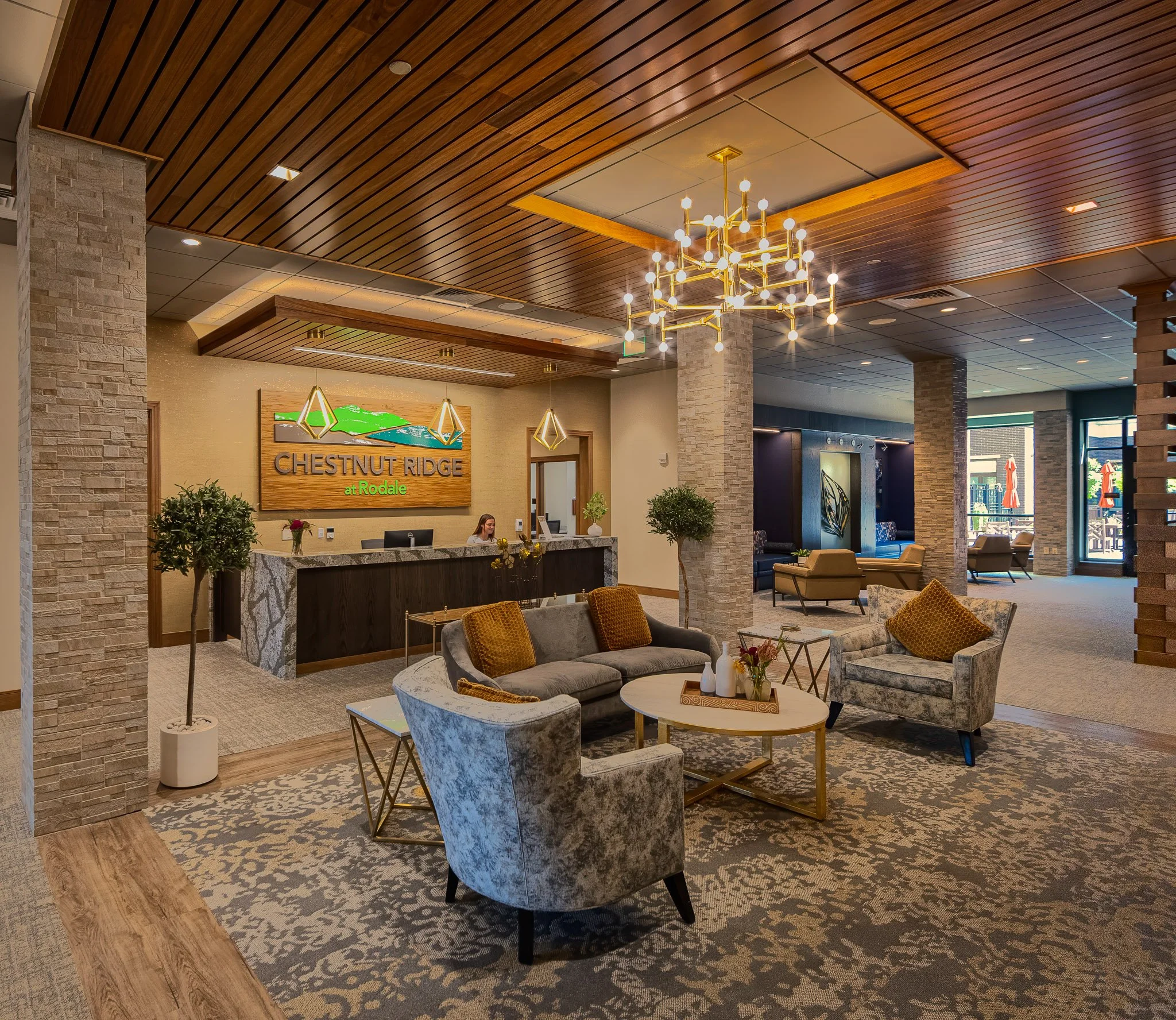 Company and Retirement Community lobby with a reception desk, seating area with sofas and armchairs, decorative plants, chandelier, and large windows.