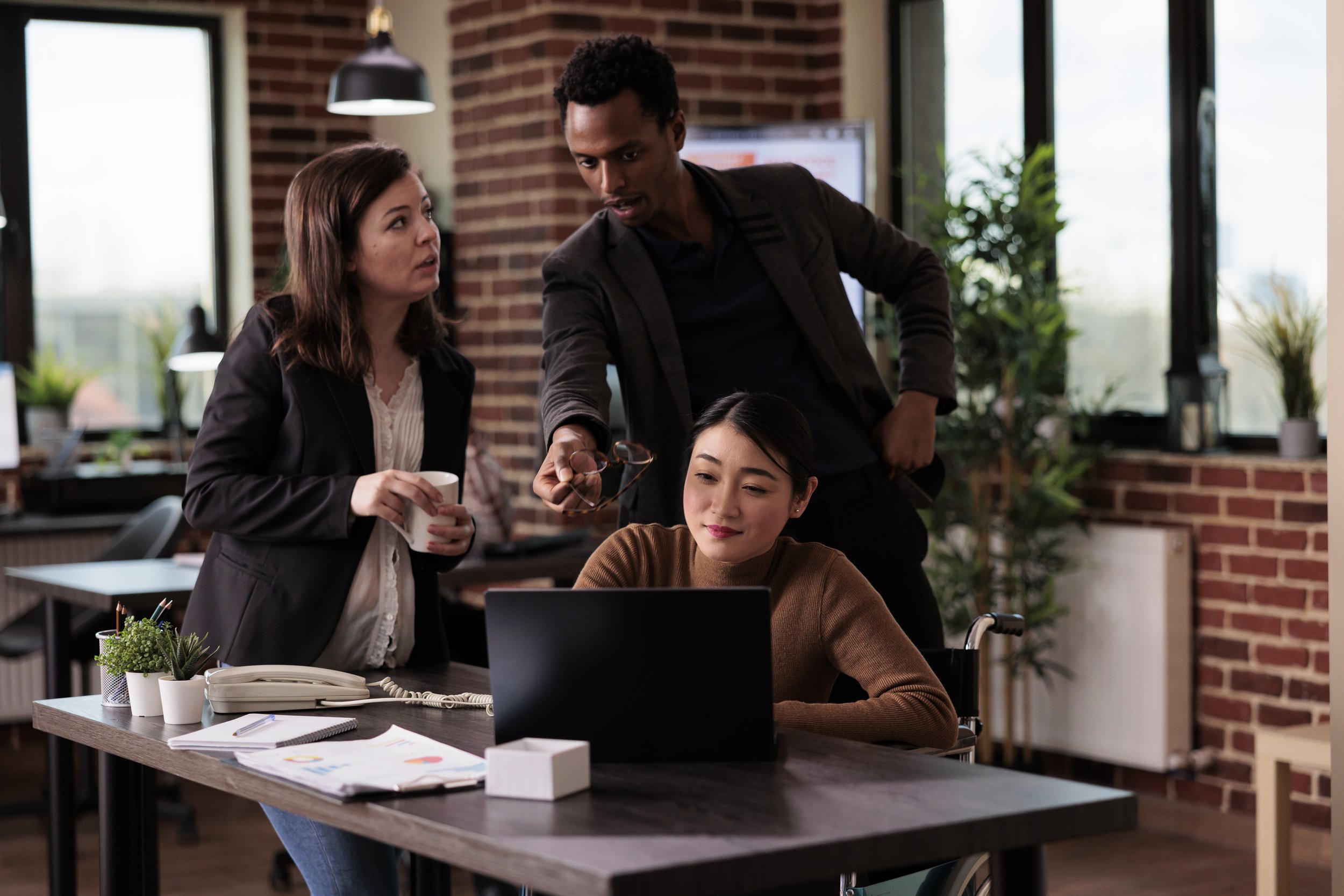 Three people in a modern office collaborating around a laptop, with one person seated and two standing, brick wall background, plants and office supplies on the table.