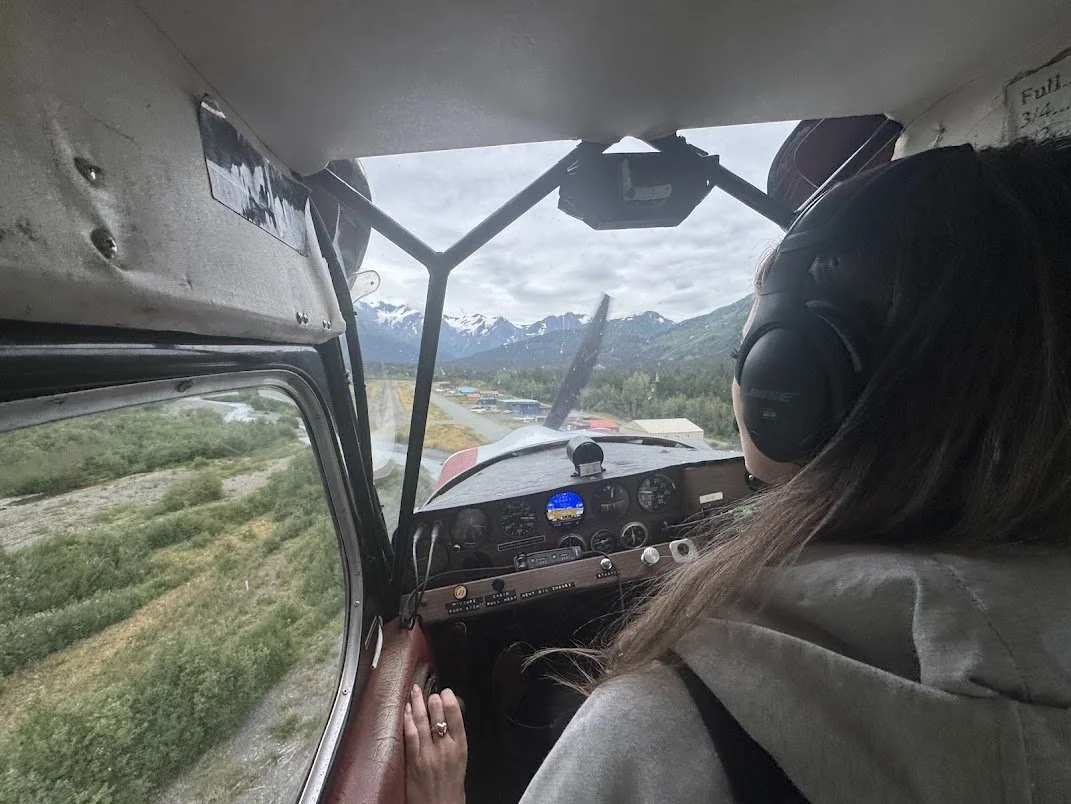 View from inside a small aircraft cockpit, with a woman wearing headphones on the right side, flying over mountains and a rural landscape with snow-capped peaks in the distance.