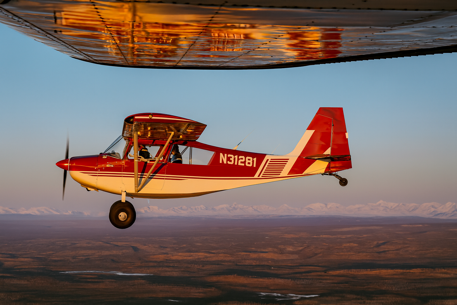 Red and white ultralight airplane flying low over a landscape during sunset, with snow-capped mountains in the background.