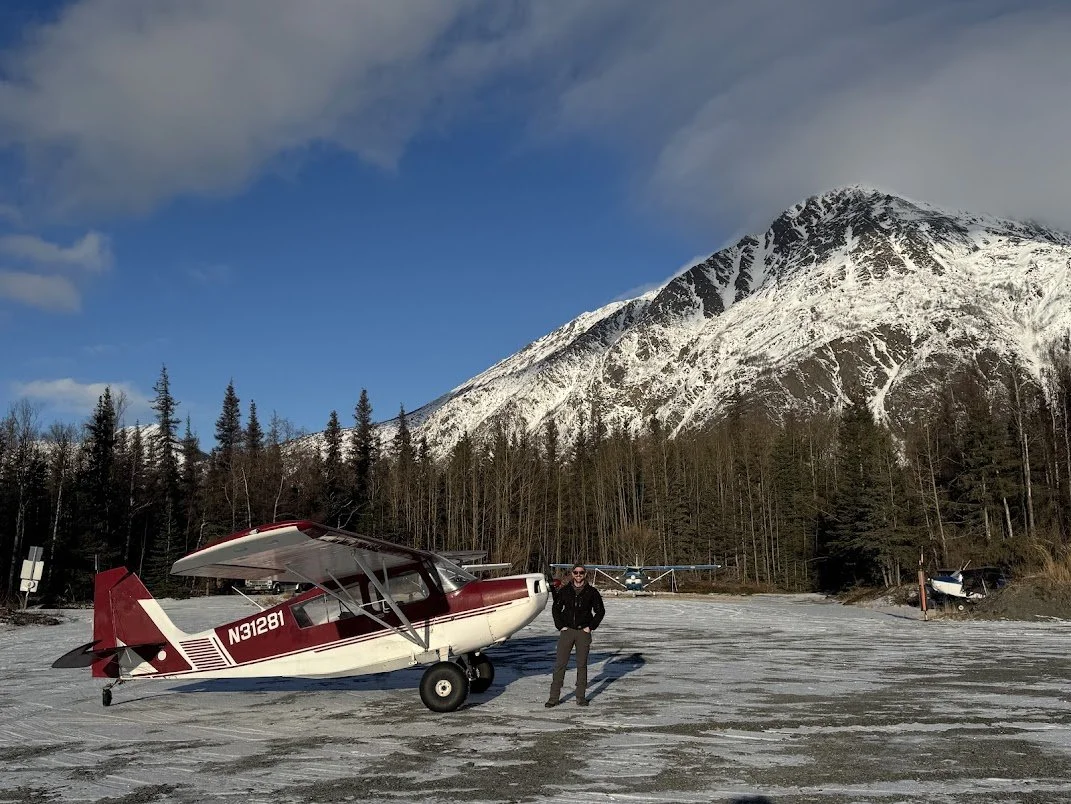 A person standing next to a small red and white airplane on a snow-covered ground, with a snowy mountain and trees in the background under partly cloudy sky.