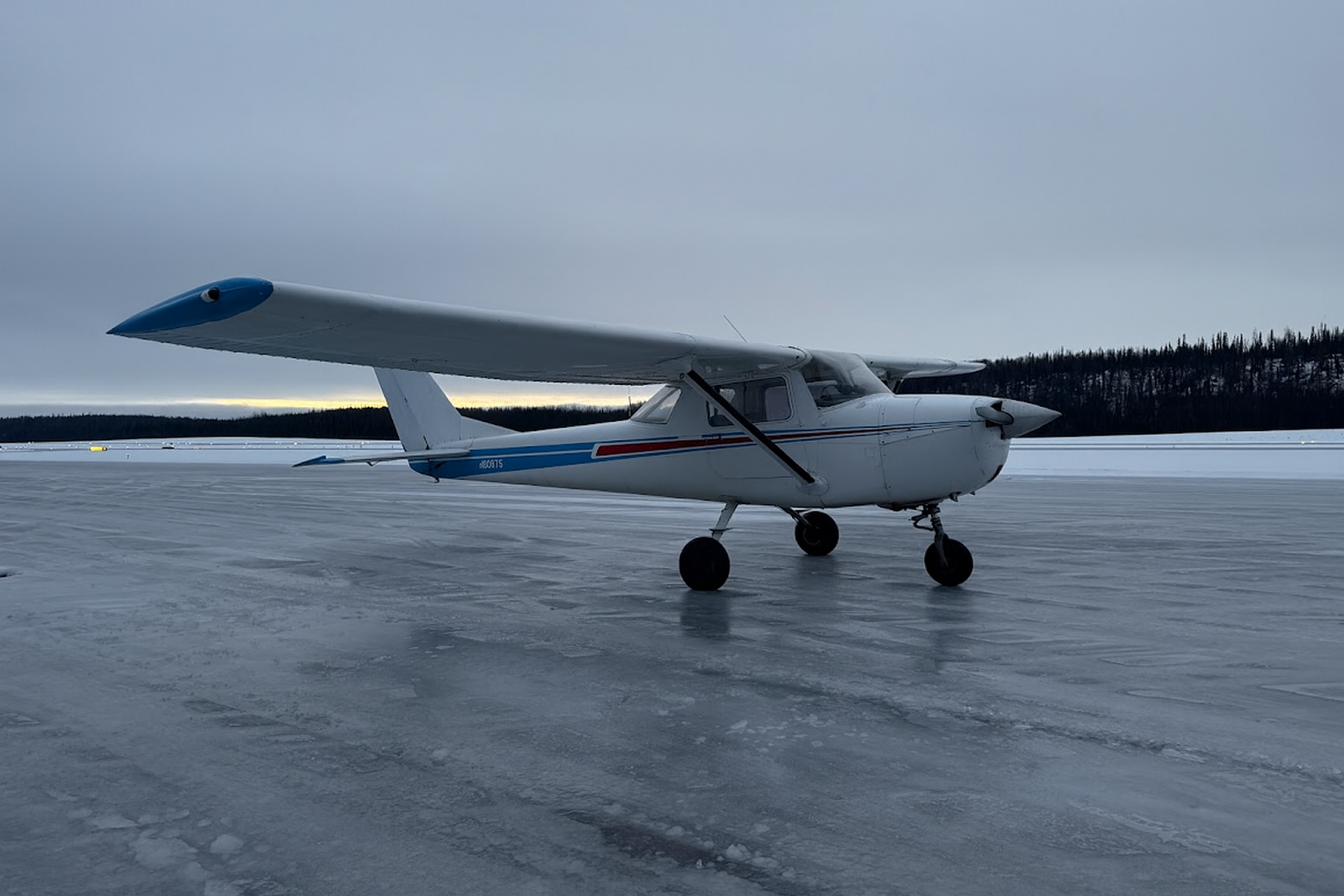 Small white airplane with blue and red stripe on icy runway with overcast sky and mountain background.