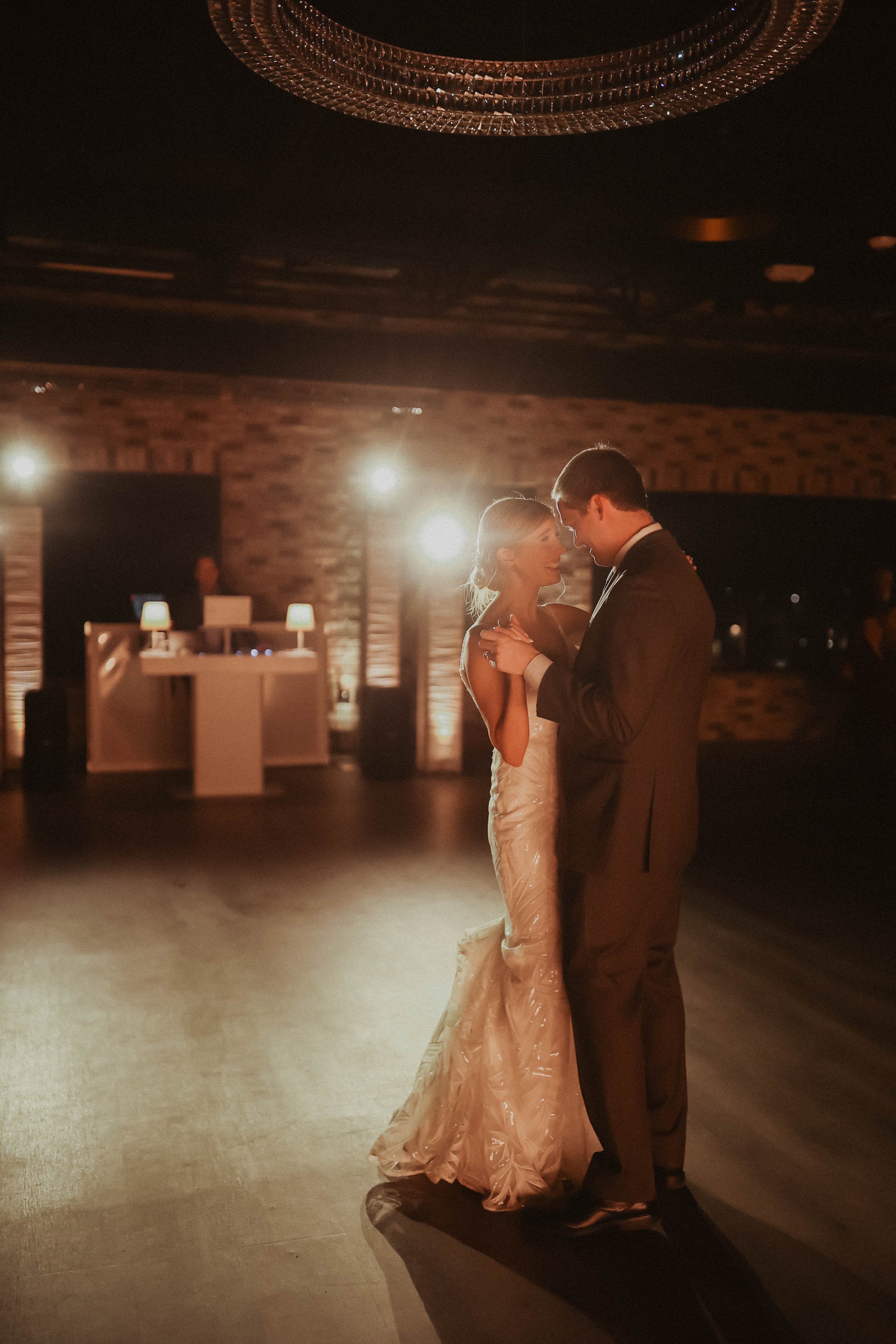 Bride and groom sharing a romantic dance on a dimly lit dance floor with light fixtures and a DJ in the background.
