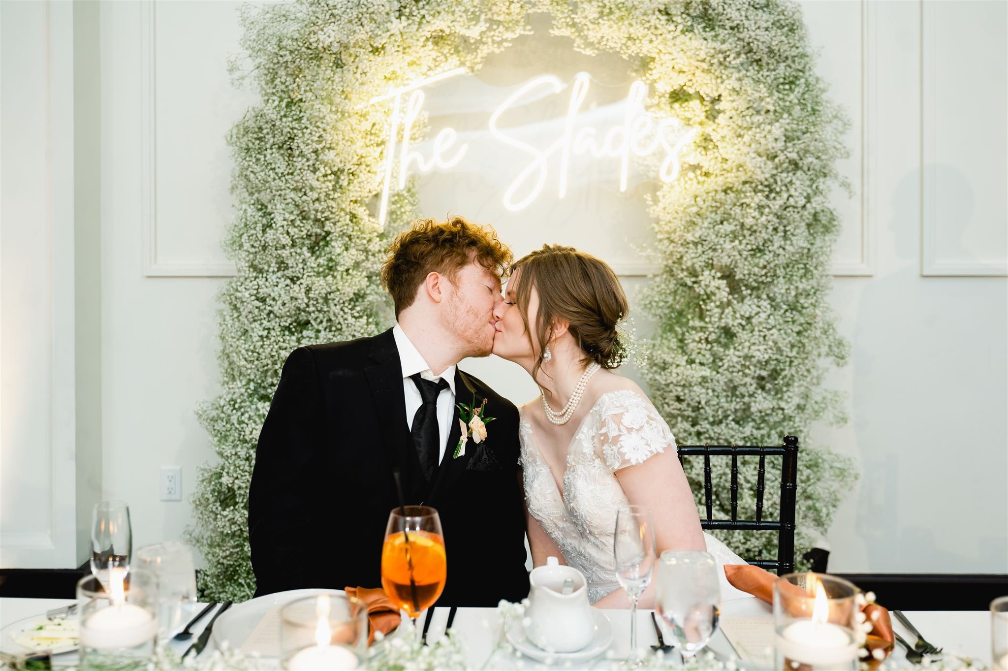Wedding couple kissing at reception table with floral backdrop and neon sign.