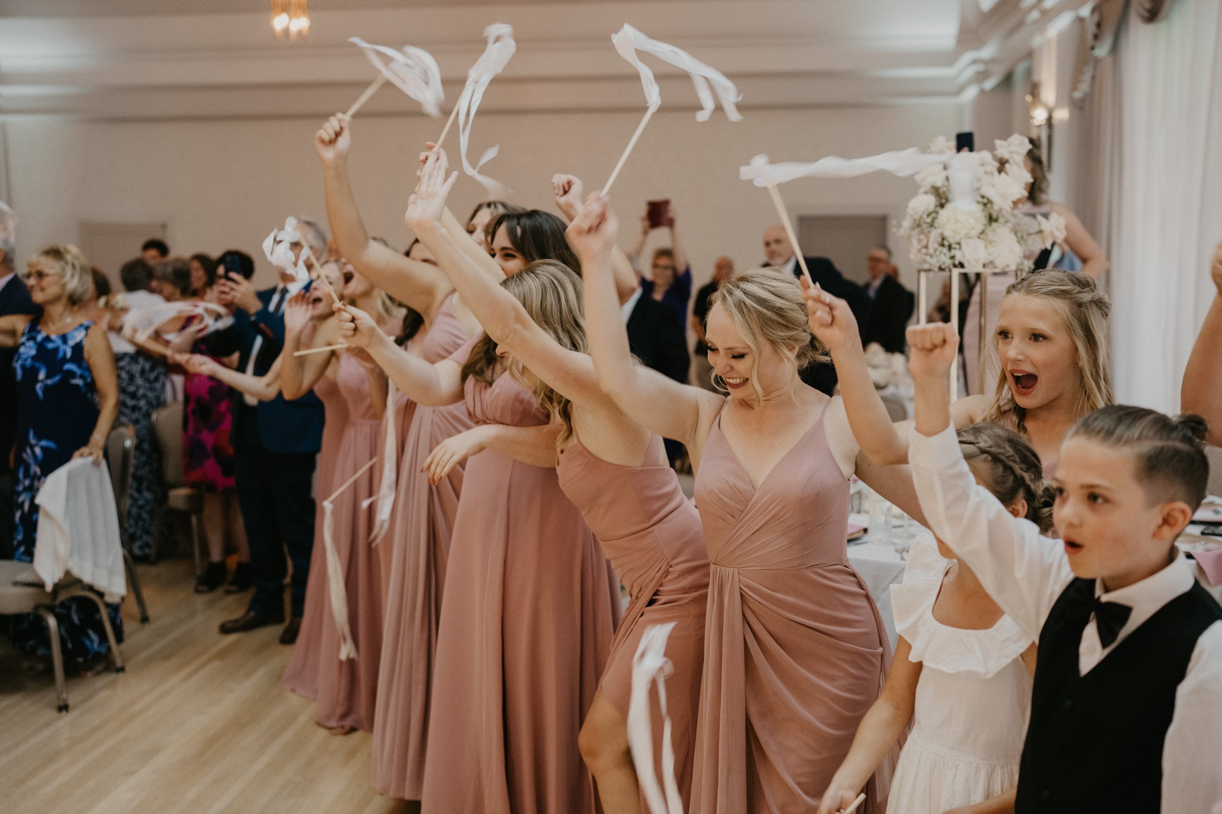 A group of people at a wedding reception, including bridesmaids in pink dresses and children in formal attire, celebrating and waving ribbon wands.