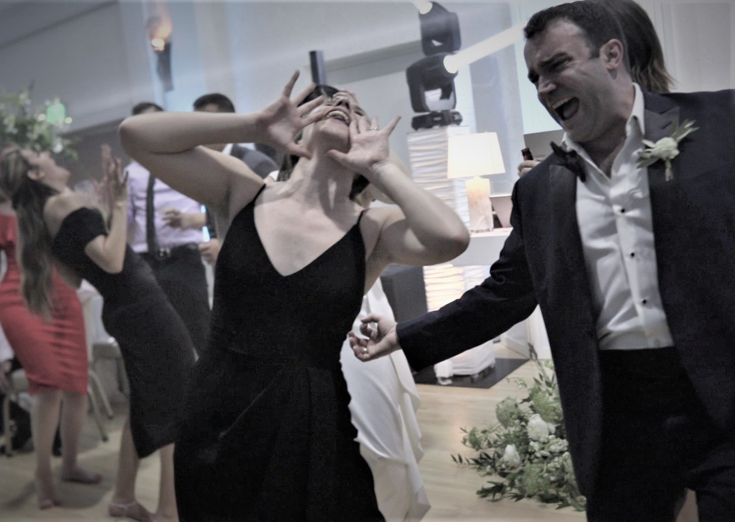 People dancing and celebrating at a wedding reception, with a woman holding a camera and a man in a tuxedo smiling and dancing.