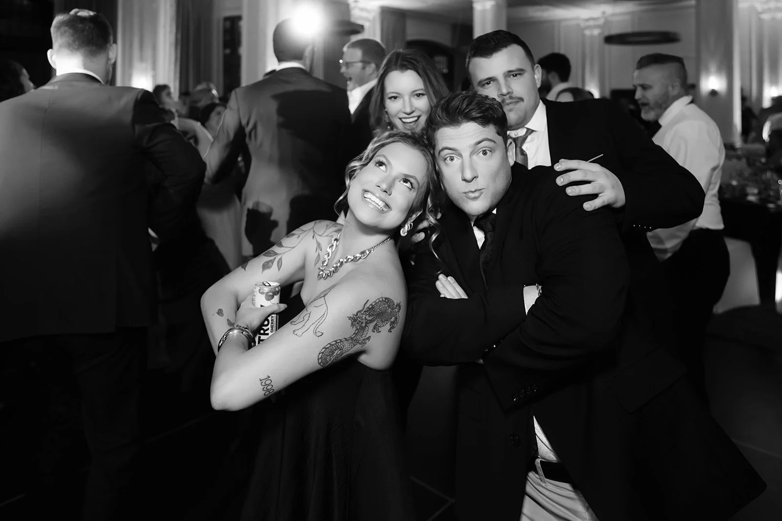 Group of people posing cheerfully at an event, with a woman holding a can and showing tattoos on her arms.