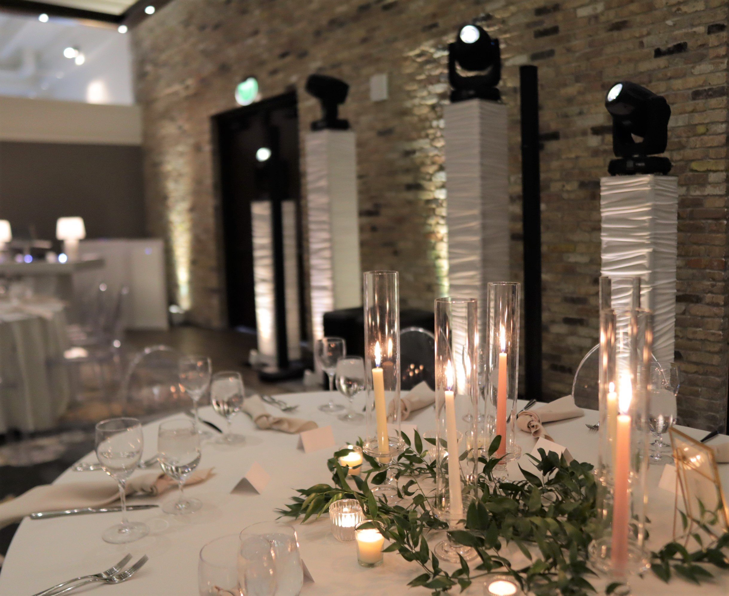 Elegant banquet table with candles, glassware, and greenery in a dimly lit event space with exposed brick walls and professional lighting equipment.