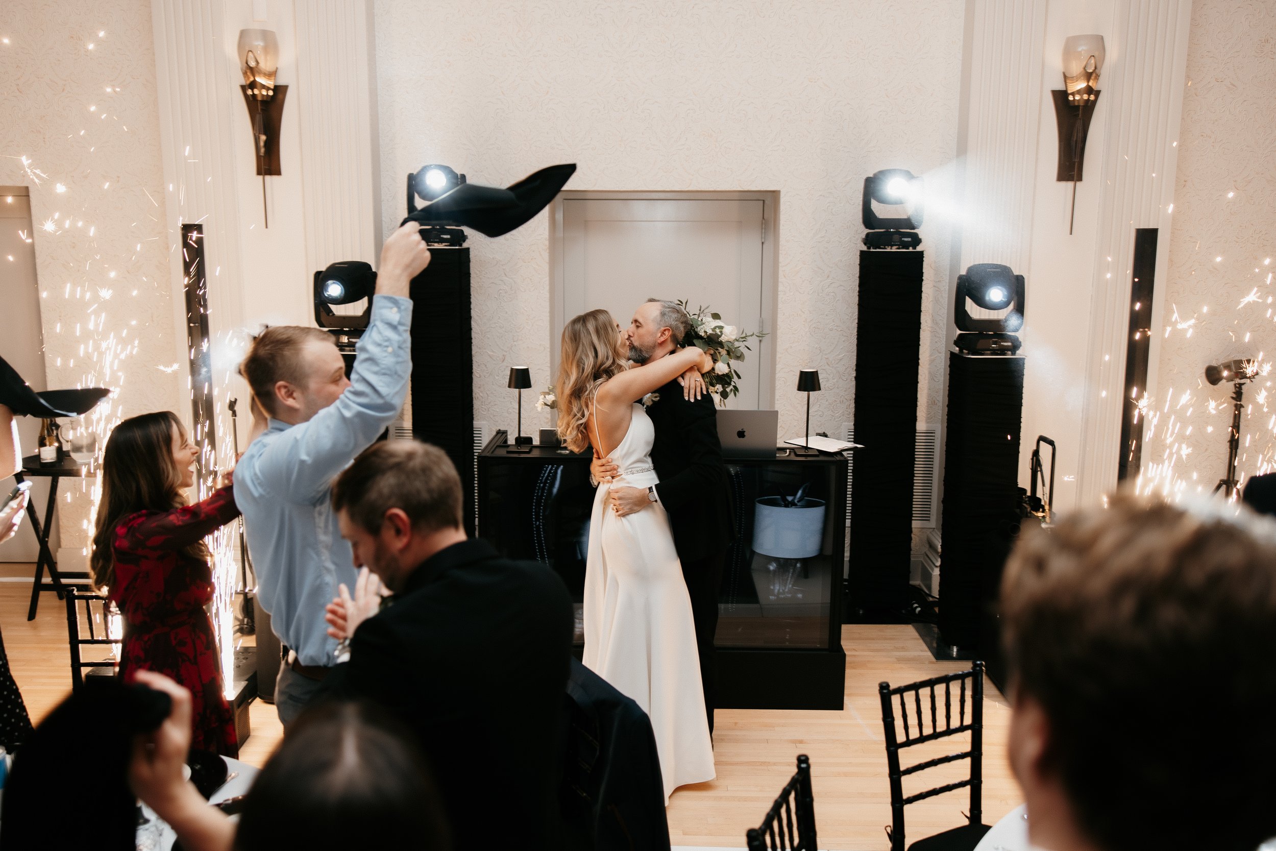 Bride and groom kissing in a wedding reception with guests celebrating and sparklers in the background.