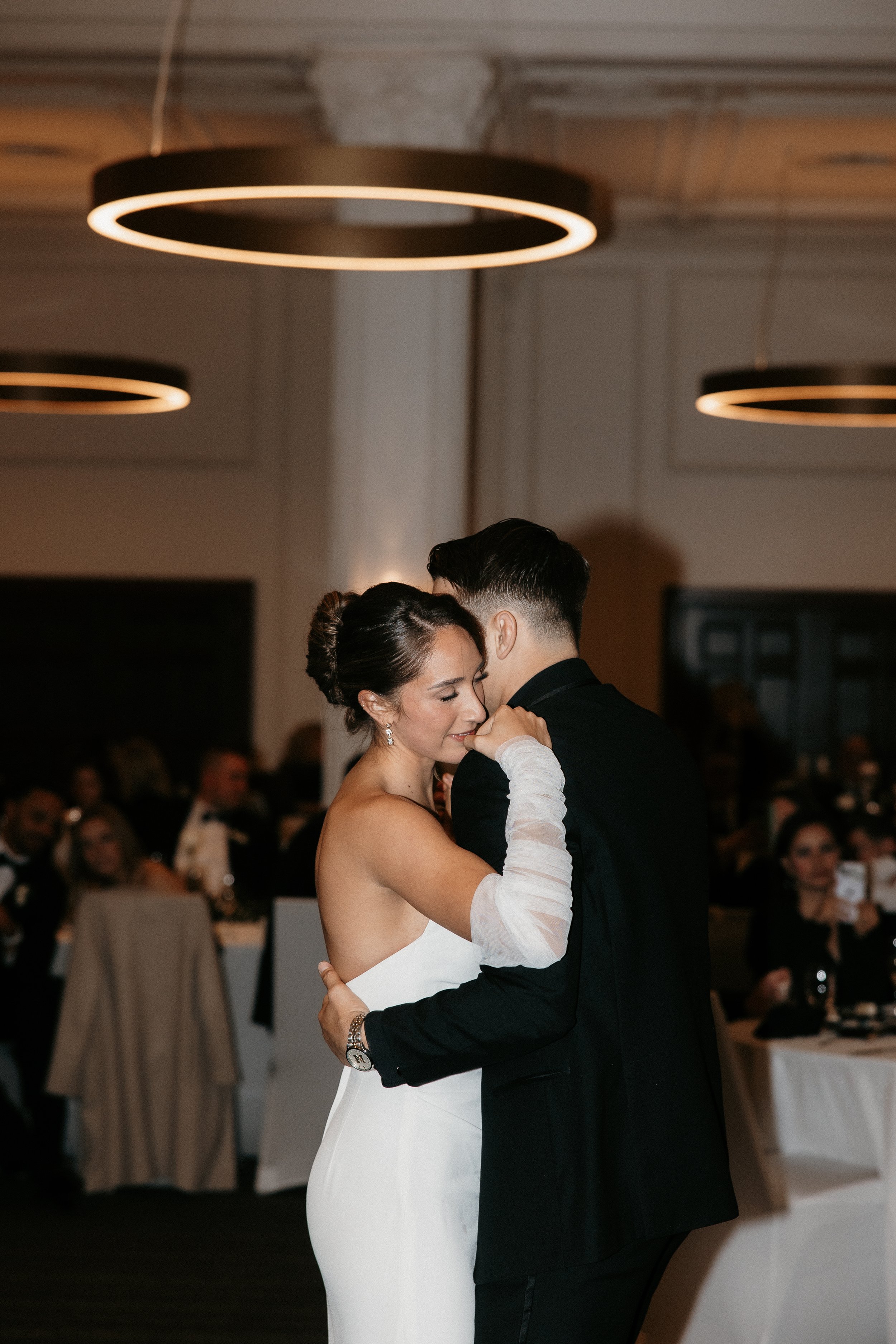 A bride and groom dancing closely at their wedding reception, with guests seated at tables in the background.