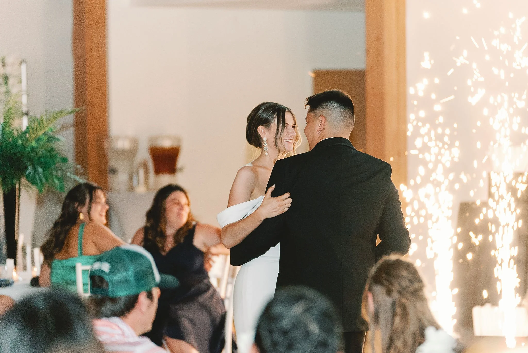 A couple dancing at a wedding reception with a sparkler effect in the background. Guests seated nearby, with drink dispensers visible in the back.