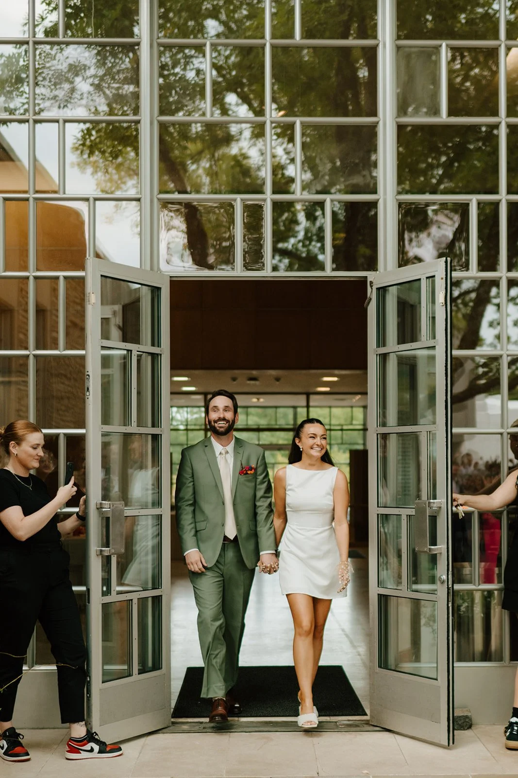 A couple walking through glass doors, holding hands, smiling; the man in a green suit and the woman in a white dress, while bystanders take photos.