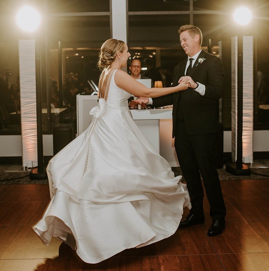 A bride and groom dancing at their wedding reception, with the bride in a white gown and the groom in a black suit, indoors with bright lighting.