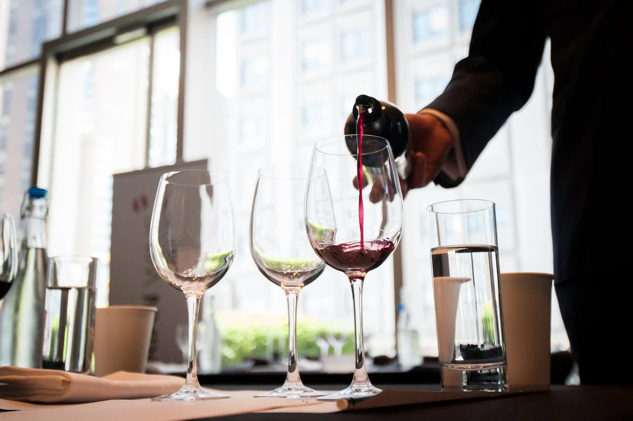A person pouring red wine into a wine glass in a bright, modern setting with large windows.